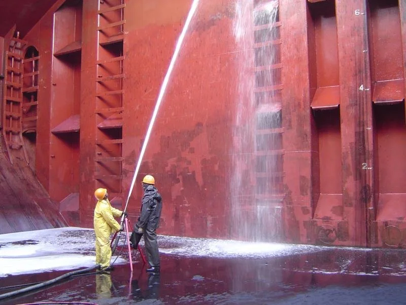 Two workers in yellow and black protective gear and helmets are cleaning the underside of a large, rust-colored ship hull with a high-pressure water hose.