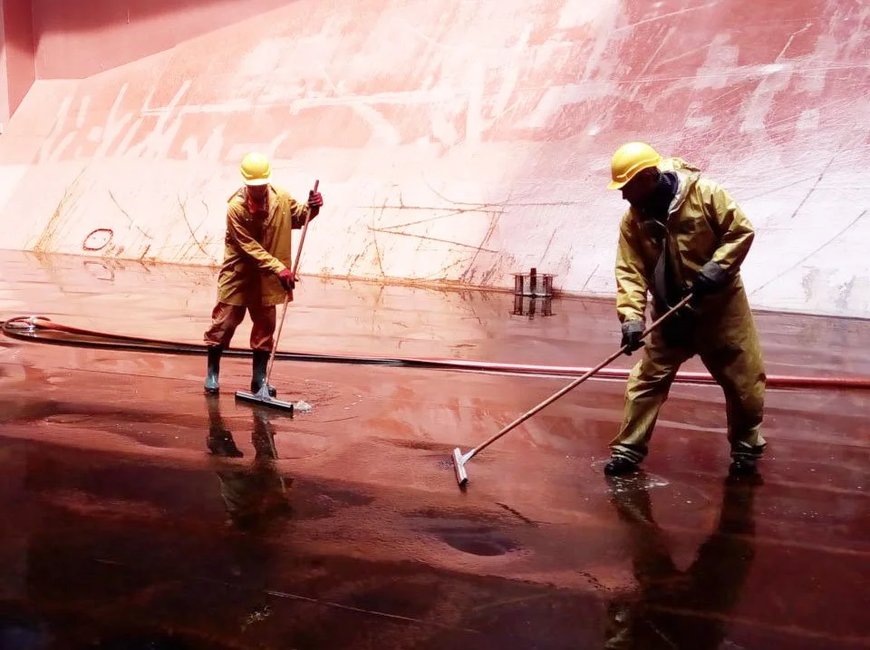 Two workers wearing yellow helmets and brown coveralls cleaning the deck of a large ship with long-handled scrub brushes.