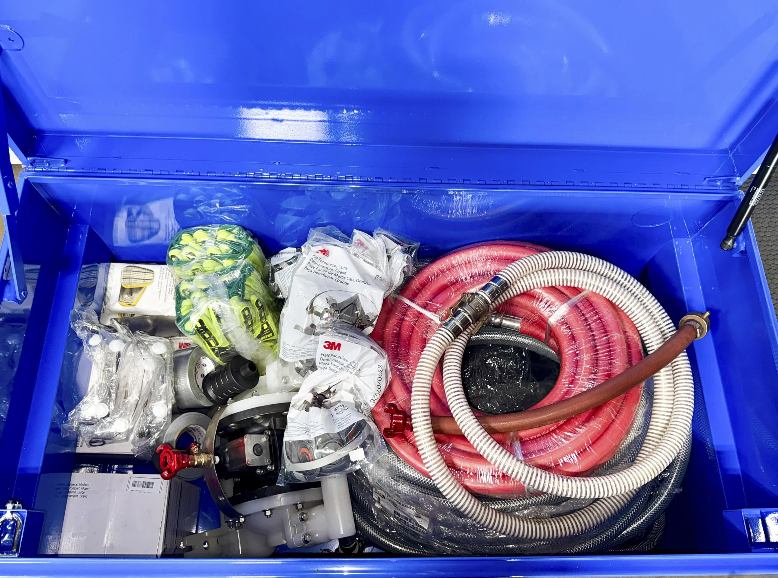 A blue plastic container filled with various hoses, connectors, and plumbing parts, including red and white flexible hoses, some plastic bags, and other small hardware items.