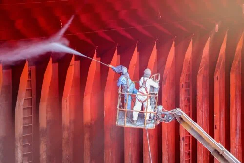Two workers in safety gear using a high-pressure water spray on a large red industrial structure.