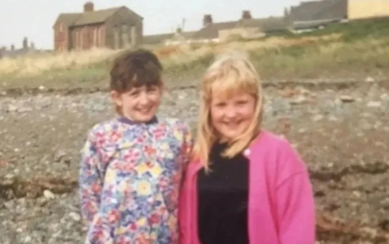 Two children, a girl and a boy, standing outdoors on a rocky area with houses in the background. The girl has blonde hair and is wearing a black top with a pink jacket, while the boy has dark hair and is wearing a colorful patterned shirt.
