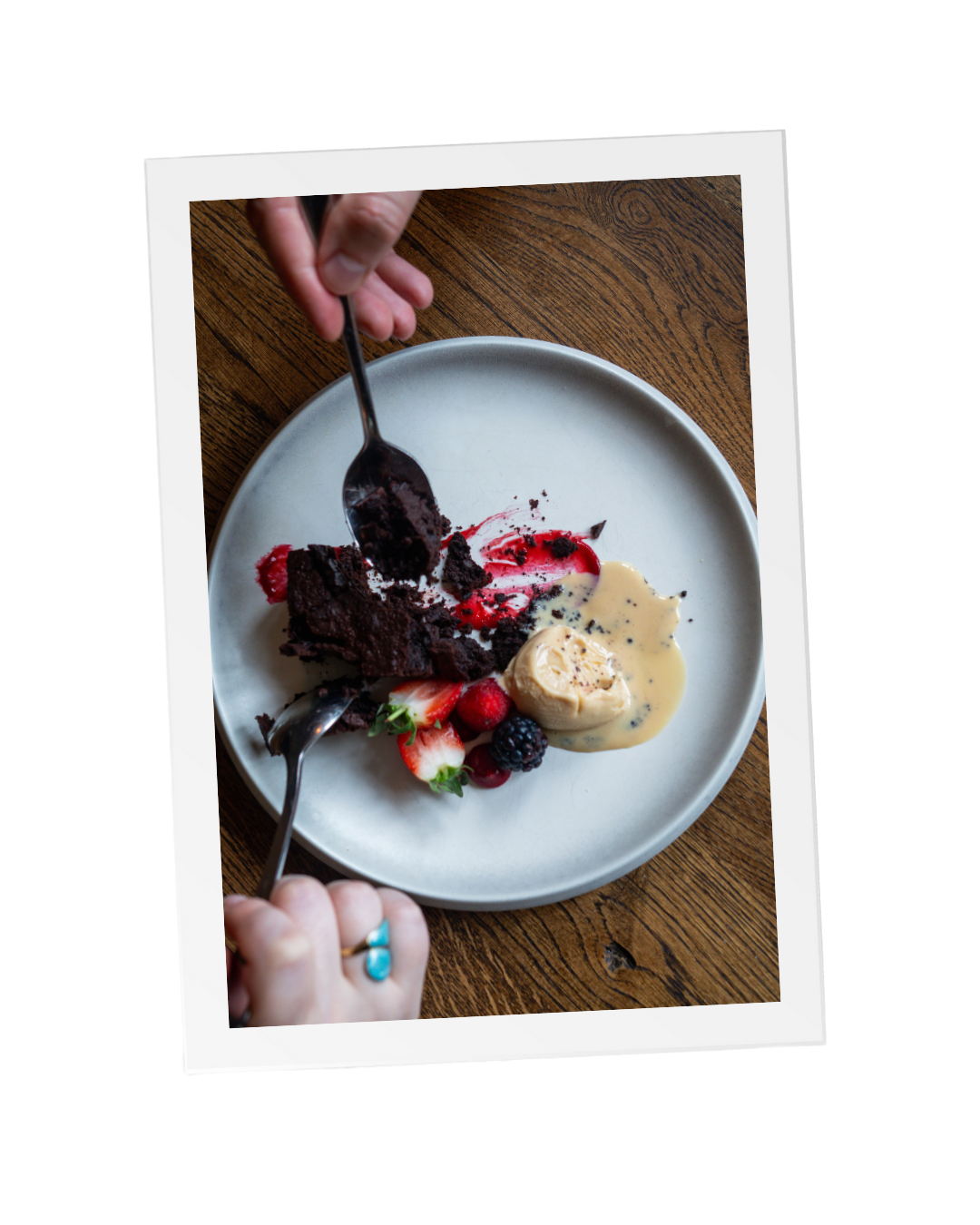 An image looking down at a wooden table with a white round plate of a delicious, gooey brownie and berries as two people enjoy the dessert for Valentine's Day at a local Sussex pub, The Thatched Inn.