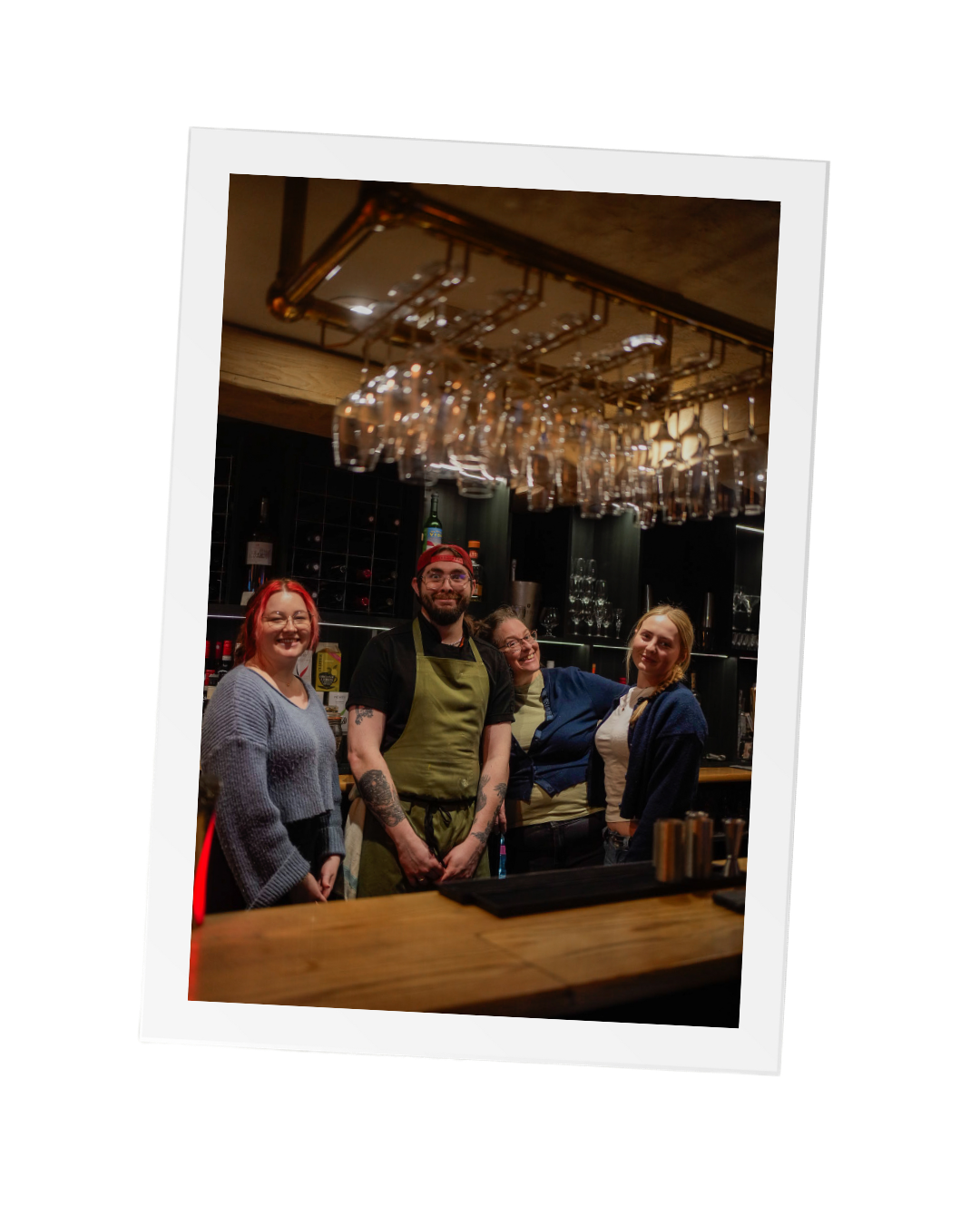 A photo of several team members smiling at the camera from behind the bar at The Thatched Inn surrounded by hanging glasses and a wide range of drink options for the local Hassocks pub.