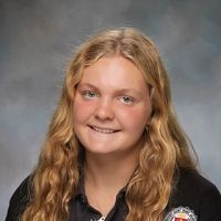 A young girl with long blonde hair smiling, wearing a black shirt with a logo on it, against a gray background.