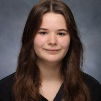 Headshot of a young woman with long brown hair, smiling subtly, wearing a dark top against a gray background.