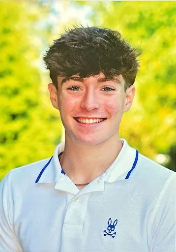 Smiling young man with dark, curly hair wearing a white collared shirt with a small bunny logo on the chest, outdoors with blurred greenery in the background.