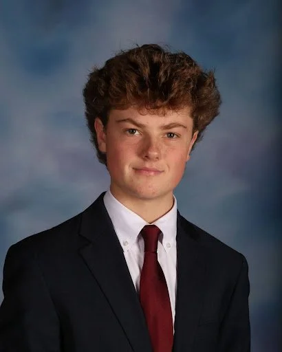 A young man with curly brown hair wearing a black suit, white shirt, and red tie, posing for a formal portrait against a blue background.