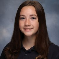 A young woman with long brown hair smiling at the camera, wearing a dark collared shirt against a gray background.