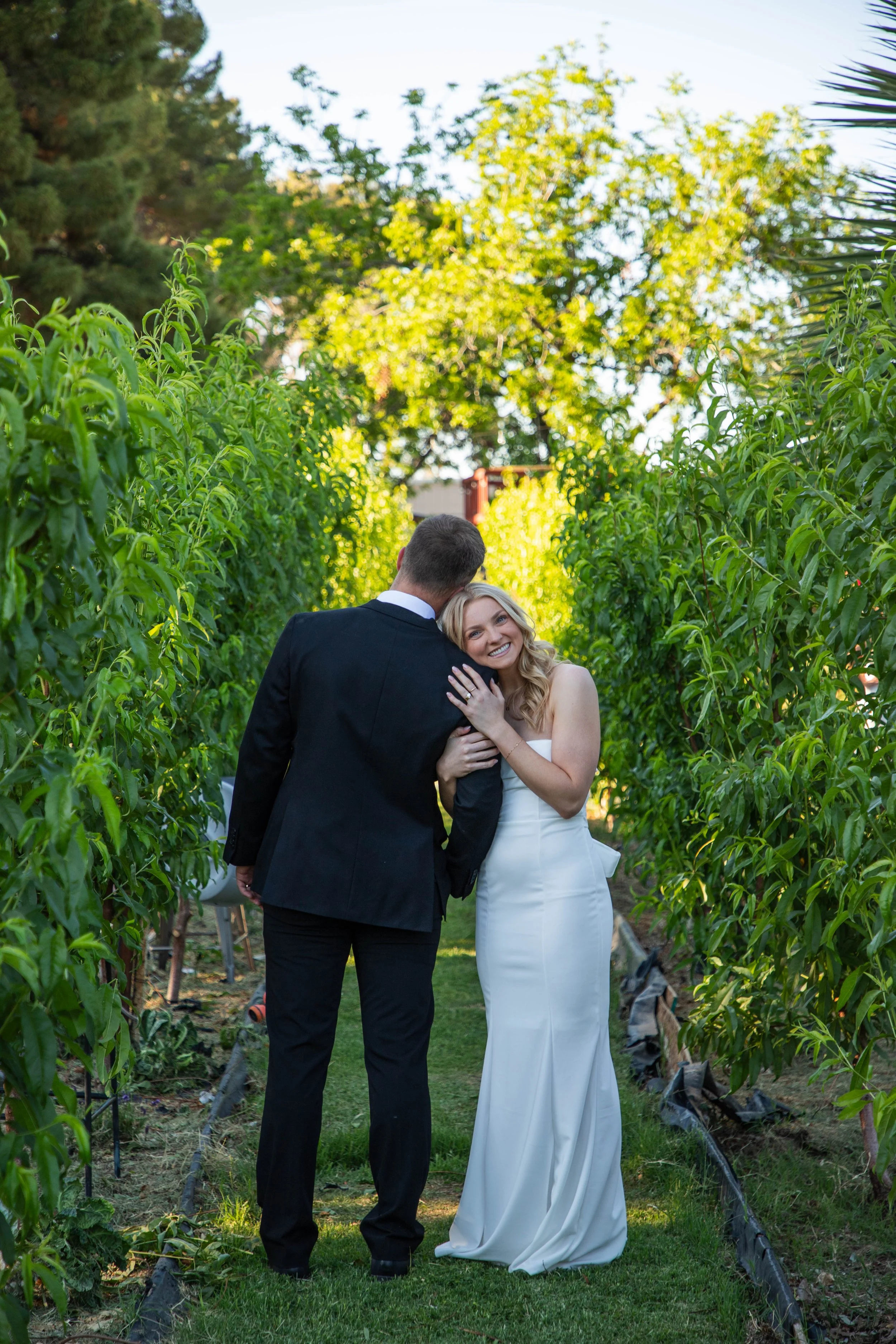 A couple dressed in wedding attire standing in a lush green orchard, smiling and embracing each other.