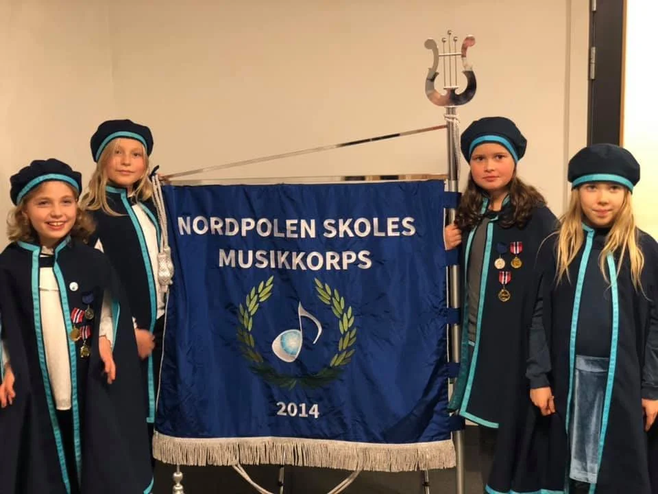 Fem barn kledd i uniformer fra Nordpolen Skoles Musikkorps står ved siden av et blått banner med inskripsjonen "NORDPOLEN SKOLES MUSIKKORPS 2014". Barna har på seg mørkeblå hatter og kapper med turkise detaljer.