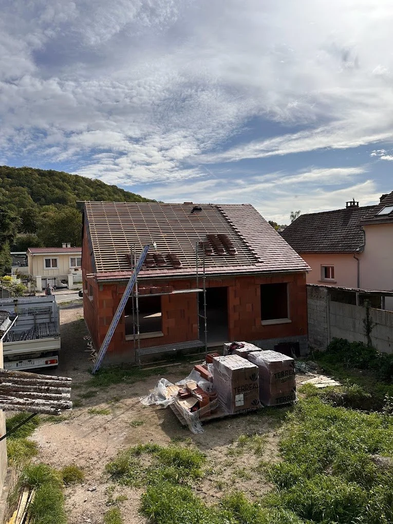 Maison en construction avec toit en tuiles, échafaudage et matériaux de construction à l'avant-plan.