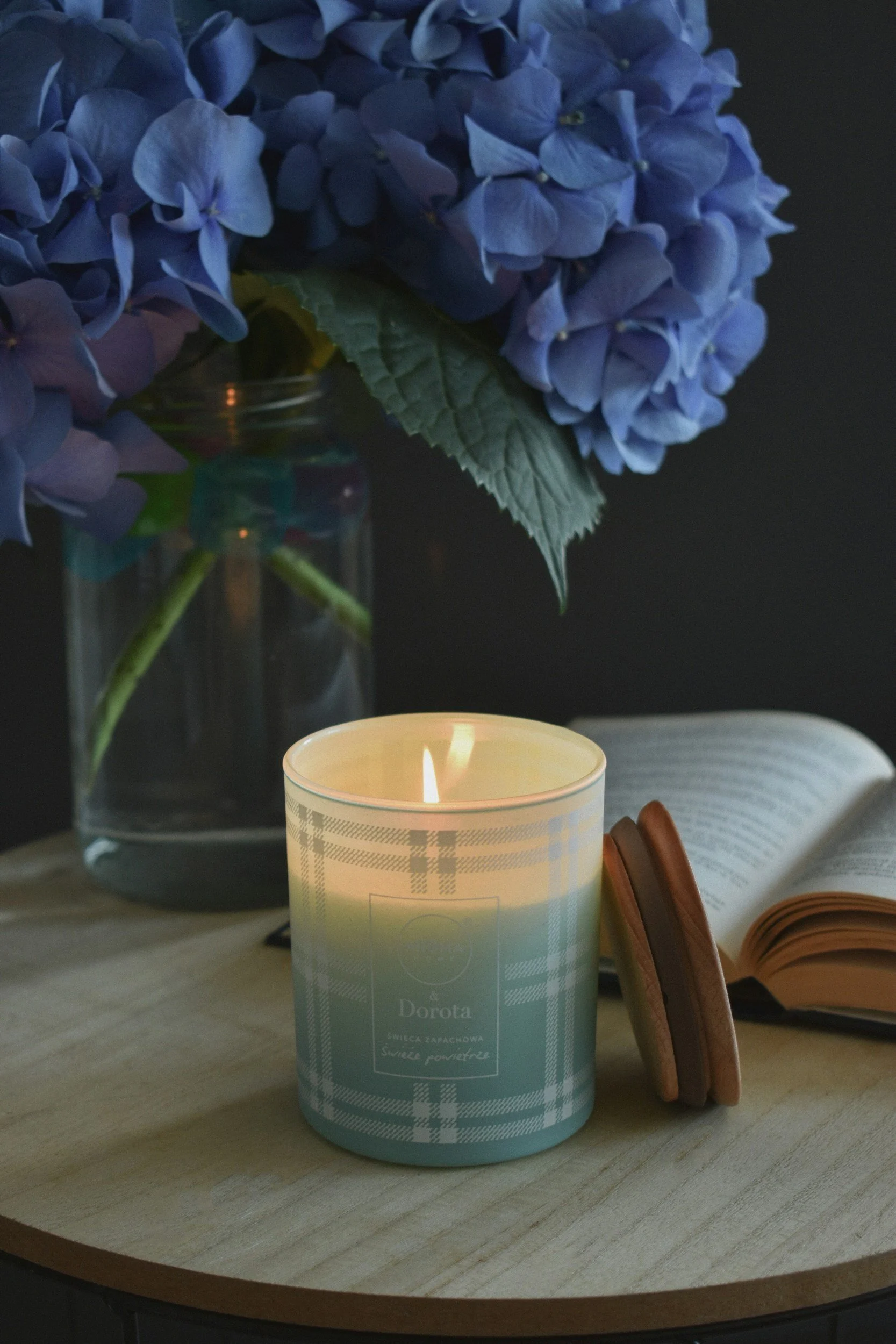 A lit scented candle with a plaid design, an open book, a pair of glasses, and a vase with purple hydrangea flowers on a wooden table.