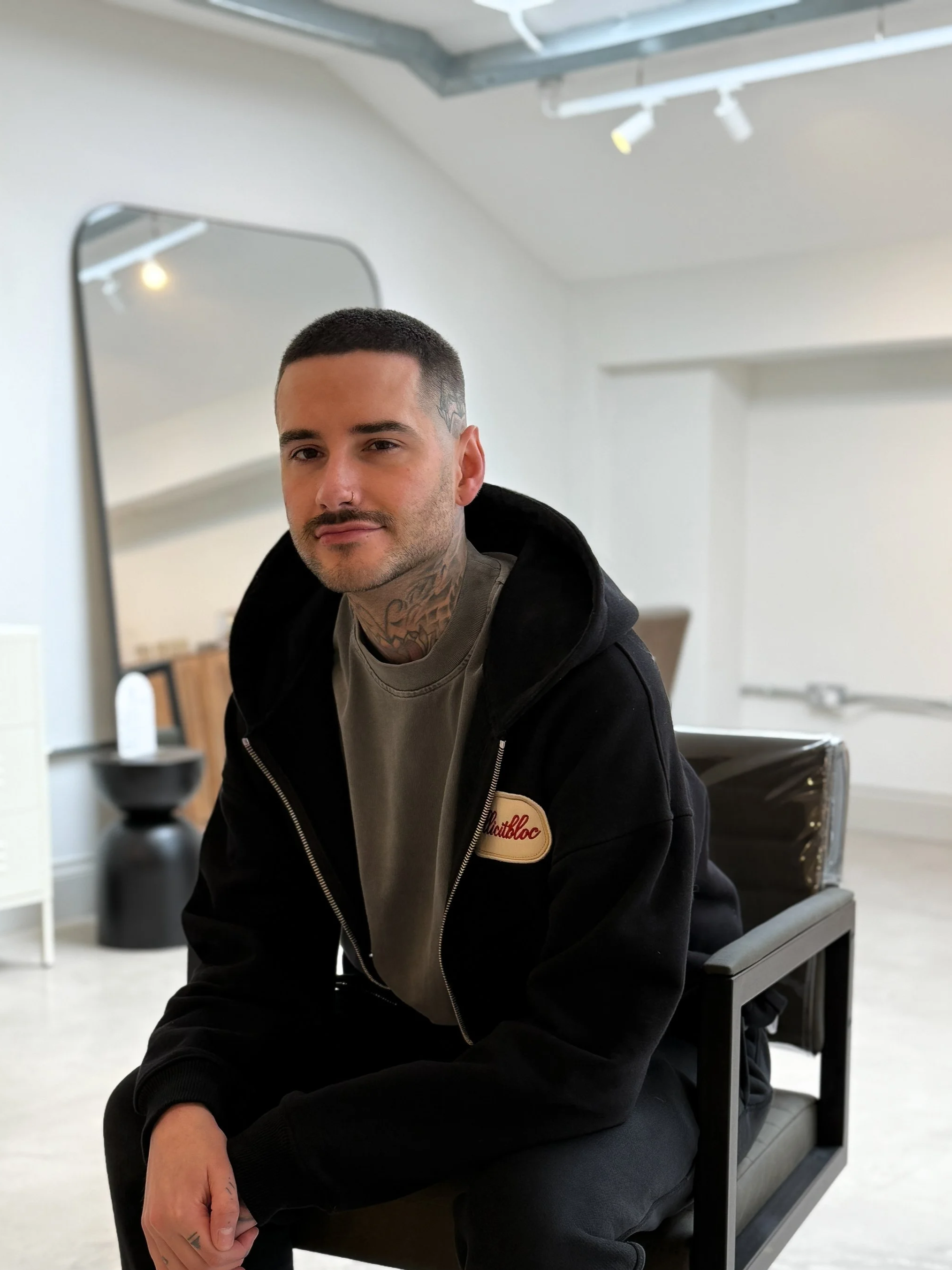 A young man with short dark hair, a nose piercing, and tattoos on his neck and fingers, wearing a gray t-shirt and a black hoodie with a patch that says 'Smithloc,' sitting on a modern black chair in a room with white walls, a mirror, and ceiling spotlights.