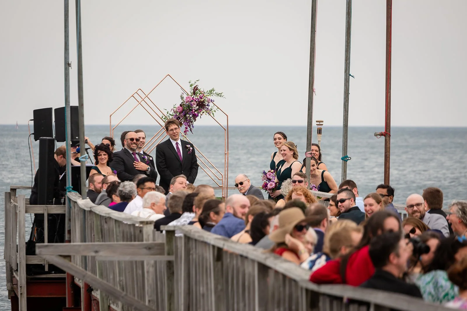 Wedding ceremony on a dock overlooking the ocean with a bride, groom, bridesmaids, groomsmen, and guests.
