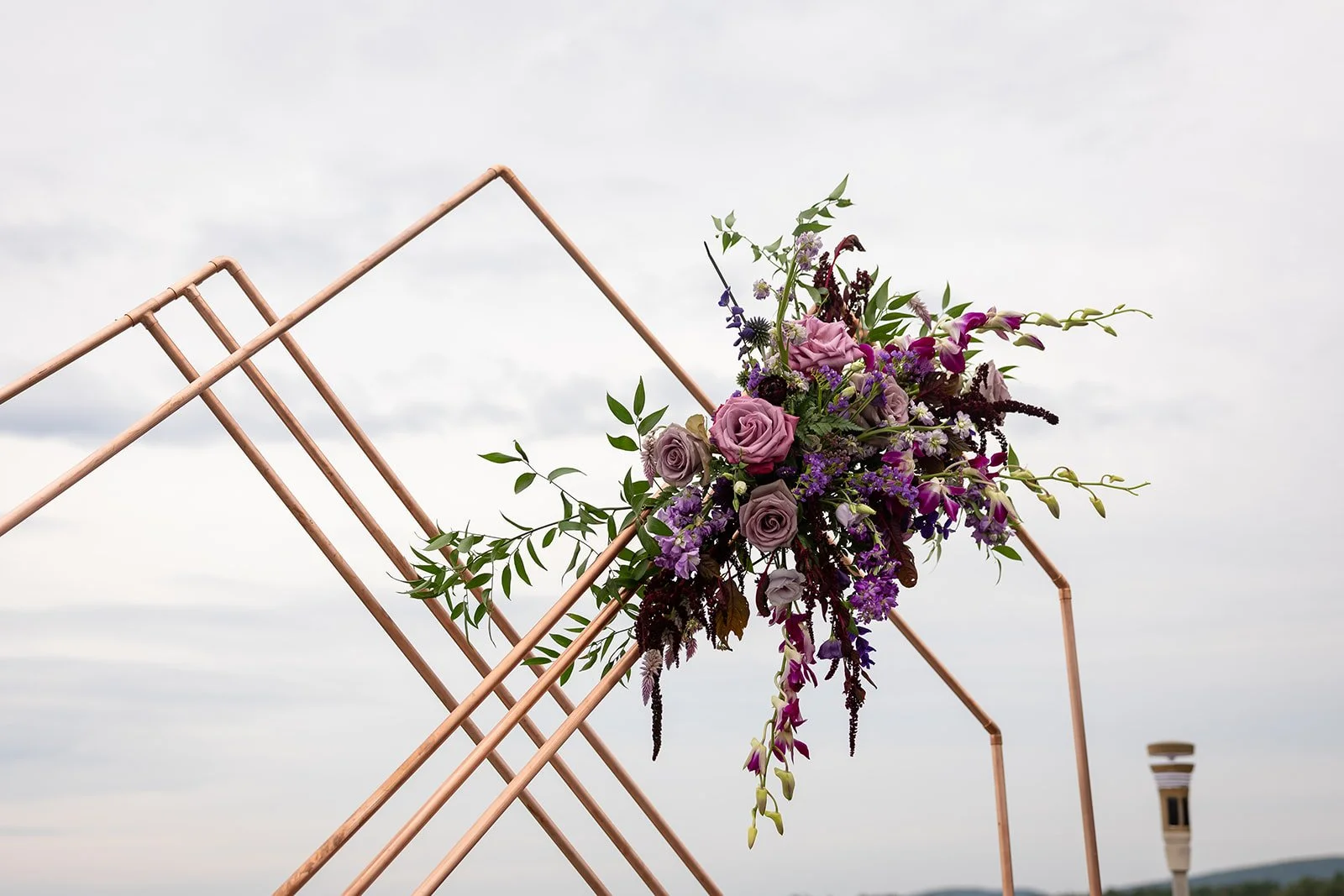 A decorative floral arrangement with purple and pink roses, purple and white orchids, and green foliage attached to a copper geometric frame outdoors against a cloudy sky.