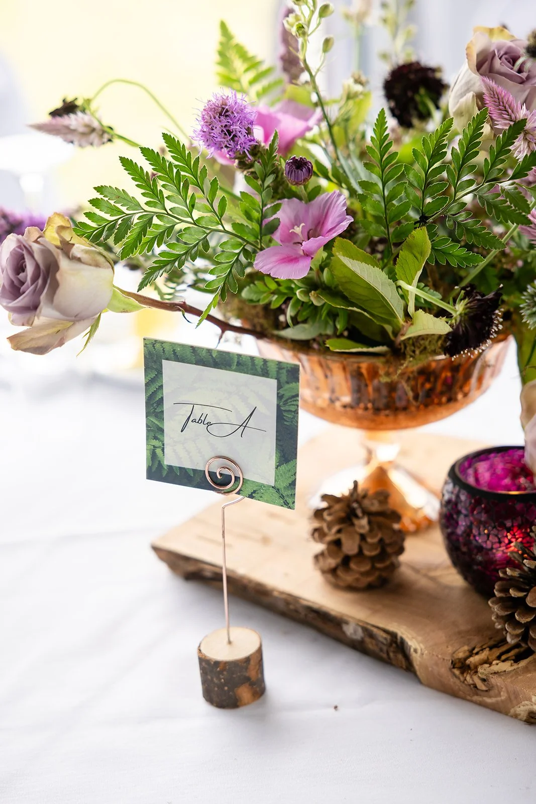 A floral centerpiece with pink, purple, and white flowers, greenery, and pinecones on a wooden base, with a table number sign reading 'Table A' attached to a small branch.