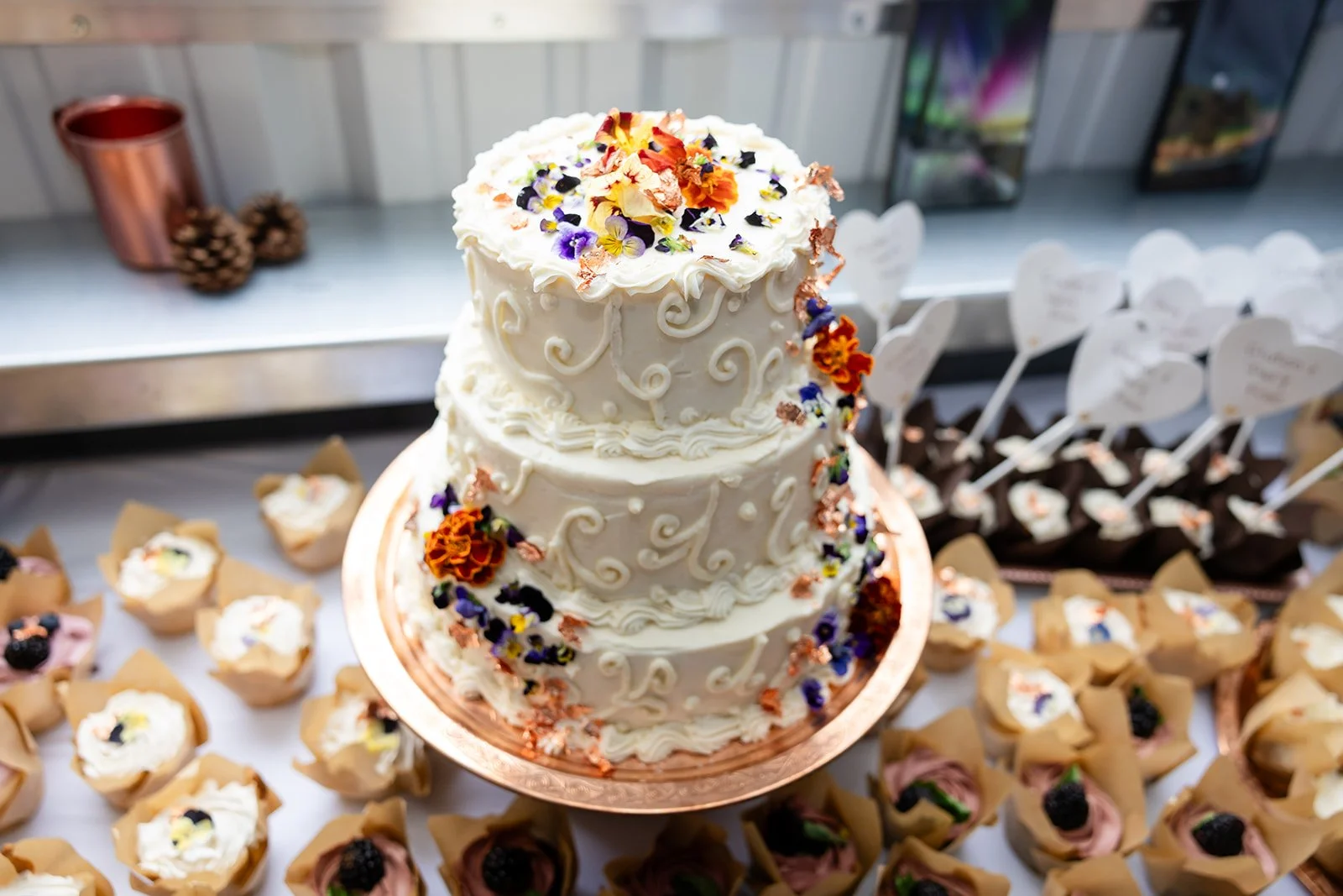 Three-tier white cake decorated with colorful edible flowers and icing swirls, displayed on a gold cake board surrounded by assorted small cupcakes with cream toppings and fruit garnishes.