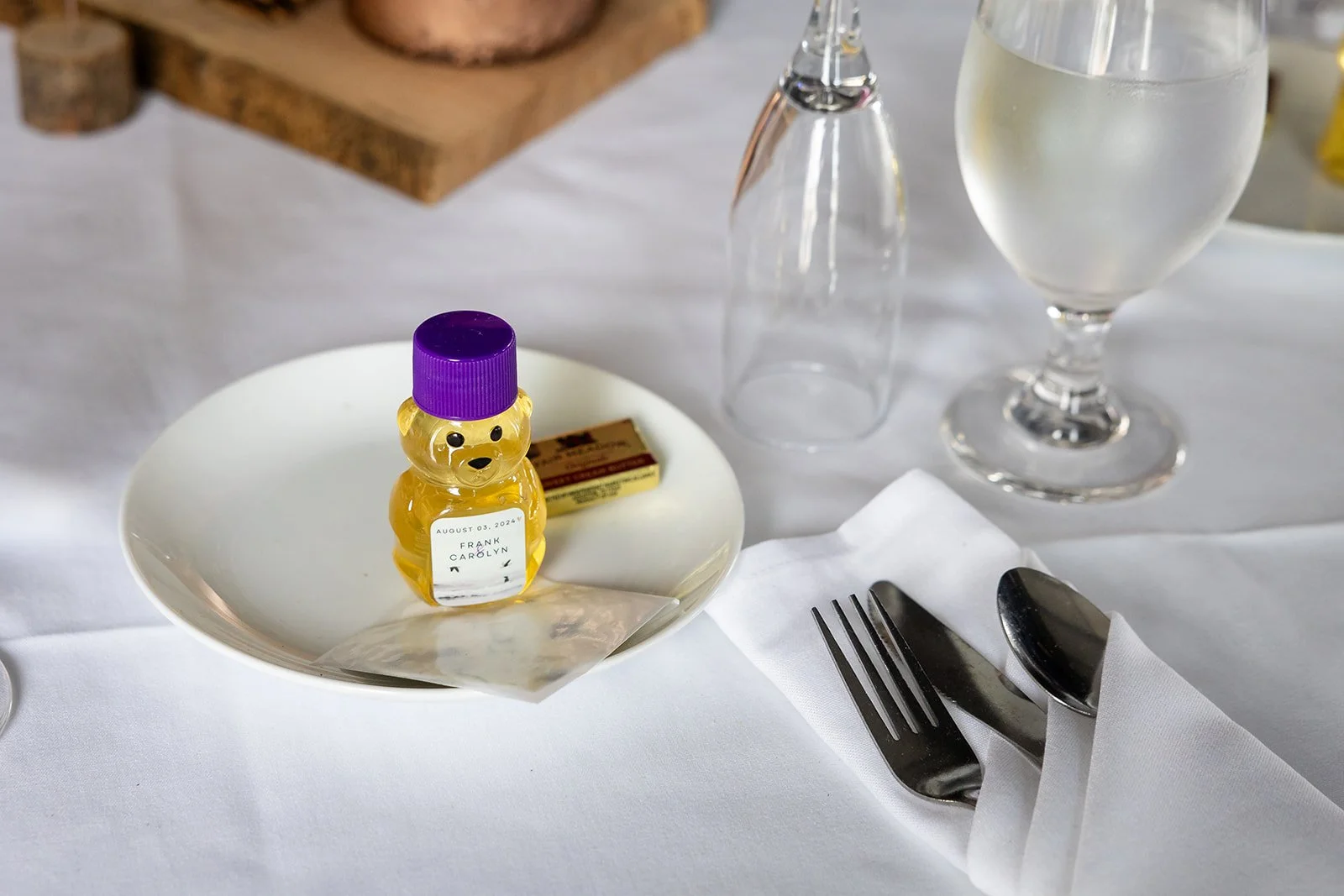 A white ceramic plate with a small bear-shaped honey jar with a purple lid, a piece of butter, a glass of water, an empty glass, and a fork and knife wrapped in a white napkin on a white tablecloth.