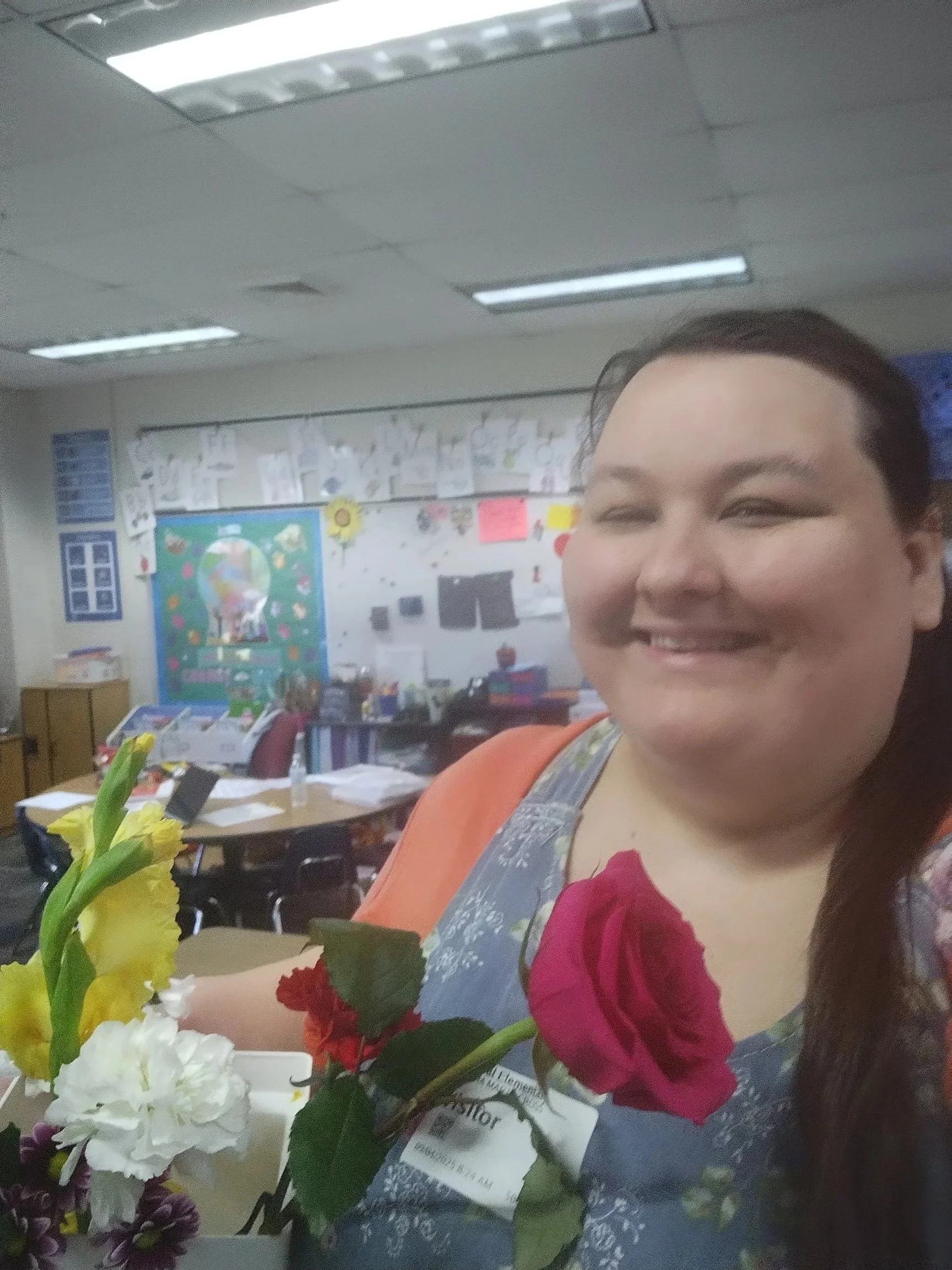 Woman smiling in classroom with colorful artwork on bulletin board, table with flowers, and fluorescent lights on ceiling.