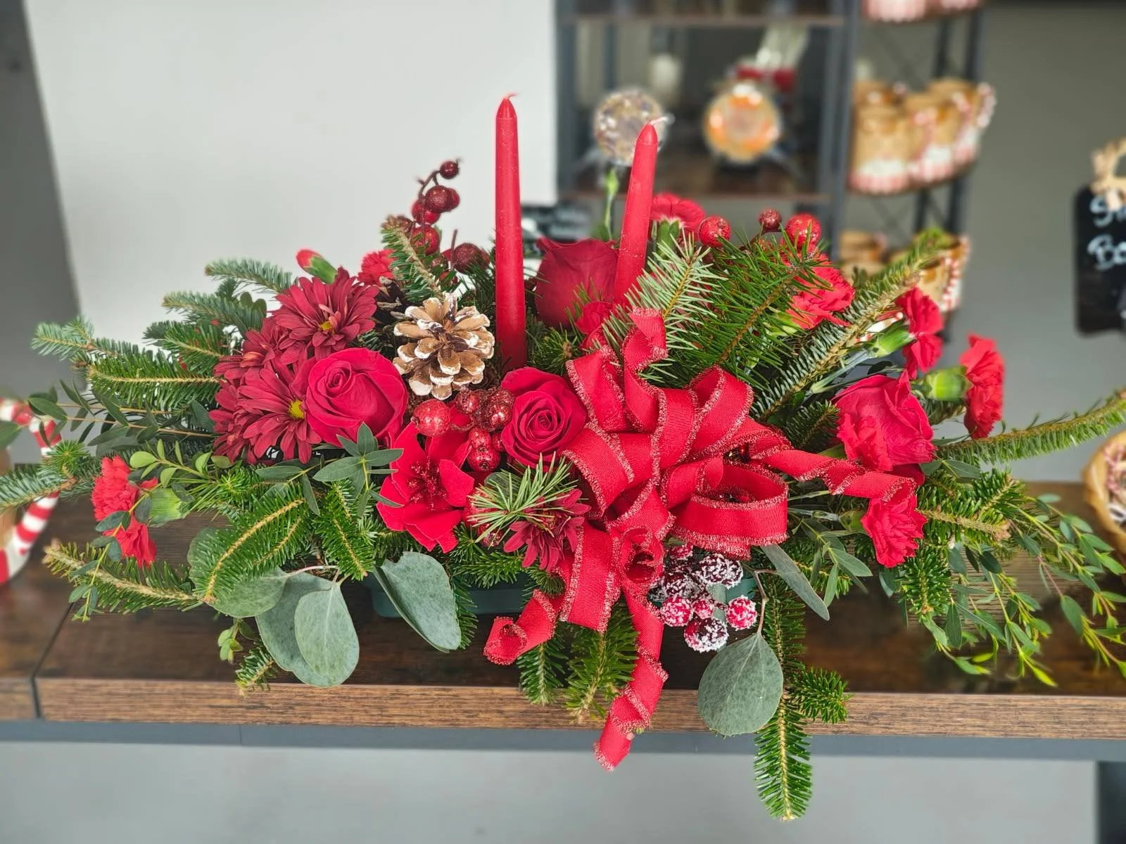 A Christmas floral arrangement with red roses, red carnations, red candles, pinecones, Christmas ornaments, green pine branches, and a red ribbon bow.