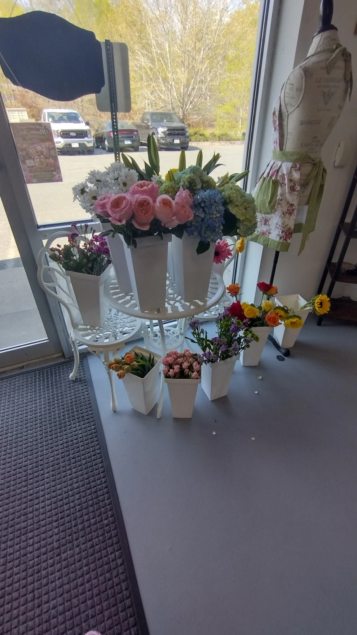 Indoor display of various colorful flowers including pink roses, white daisies, blue hydrangeas, and purple tulips arranged in white pots and a white metal table near a window, with vehicles parked outside visible through the window.