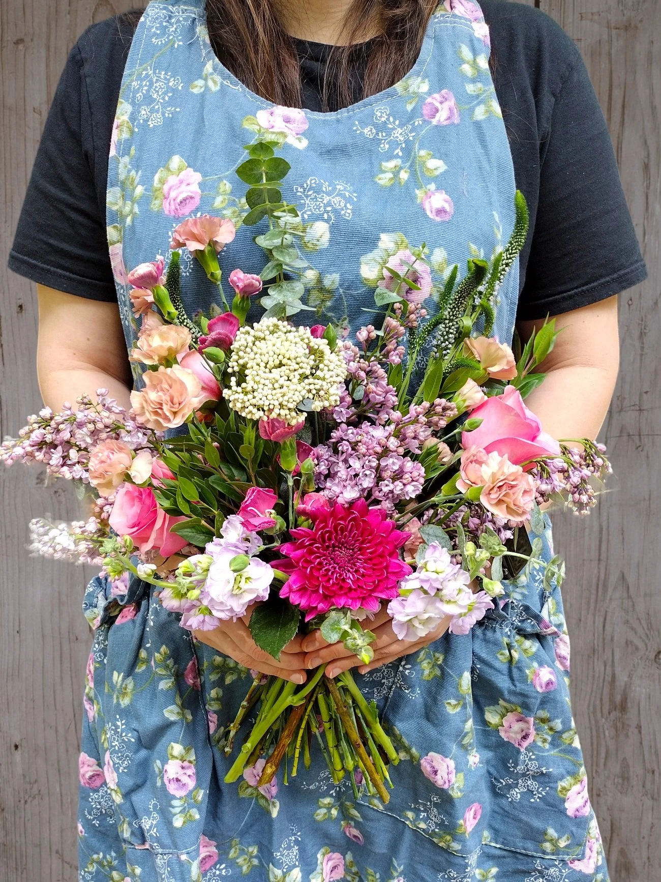 Person holding a bouquet of colorful flowers, wearing a blue floral apron.