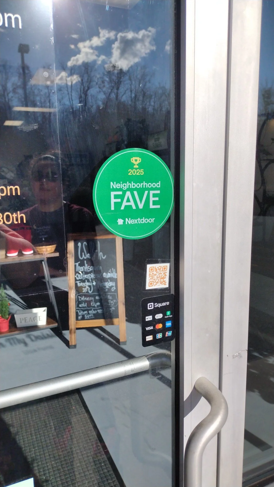 Green sticker on glass door reading '2025 Neighborhood FAVE Nextdoor' with a trophy icon. Reflection of sky with clouds and trees, and part of a person’s face taking the photo.