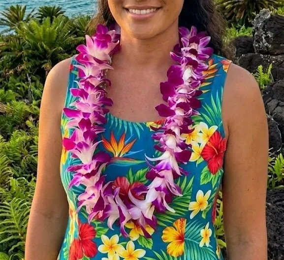 Woman wearing a tropical dress and a purple flower lei, standing outdoors with lush greenery and volcanic rocks.