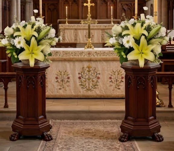 Two wooden pedestals with floral arrangements of white lilies and daisies in front of an altar with a gold cross and lit candles, in a church setting.