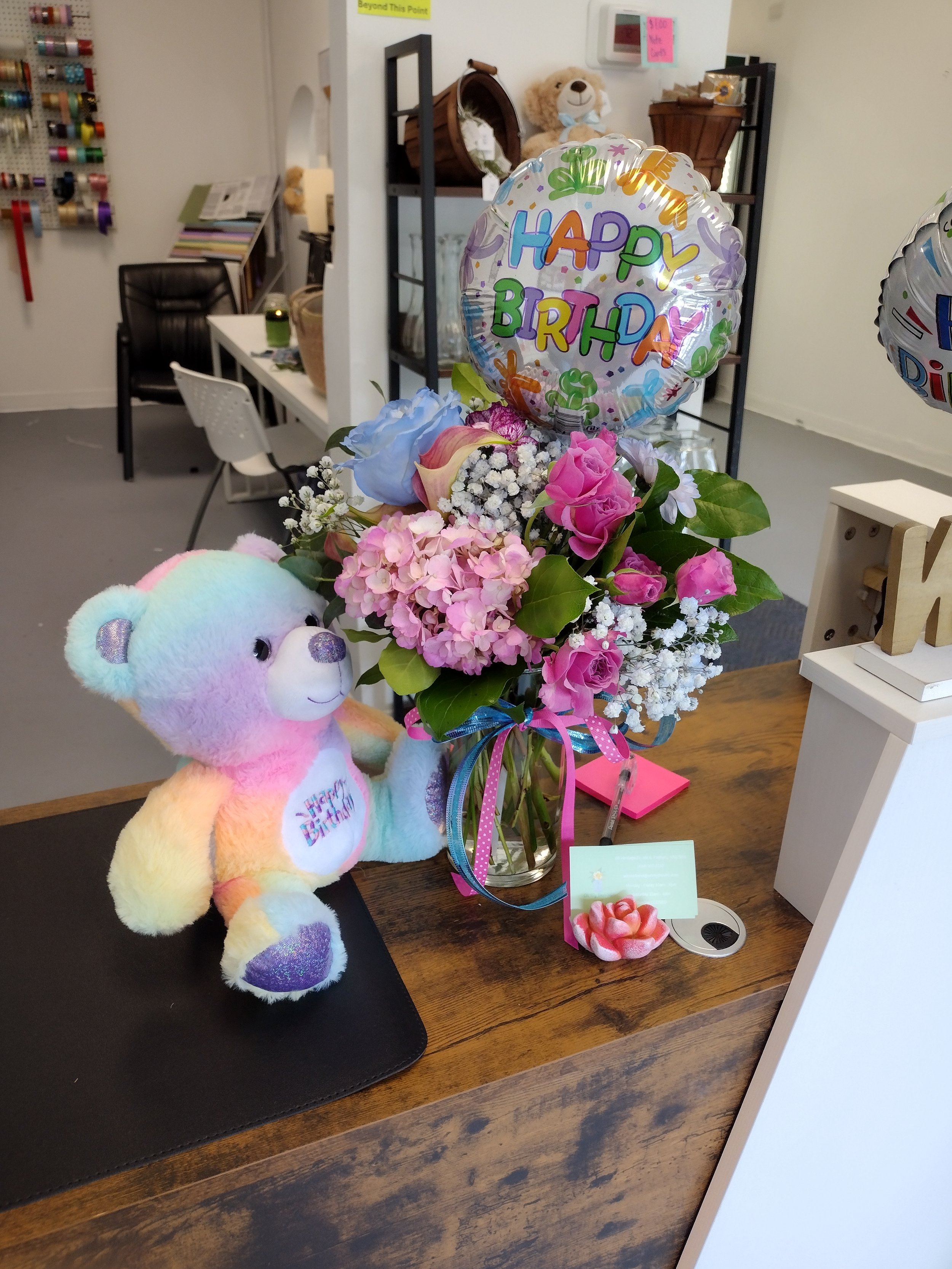 Colorful plush unicorn toy, a bouquet of pink, purple, and white flowers, and a 'Happy Birthday' balloon on a wooden table. The background shows a shelving unit, a teddy bear, and various decorations.