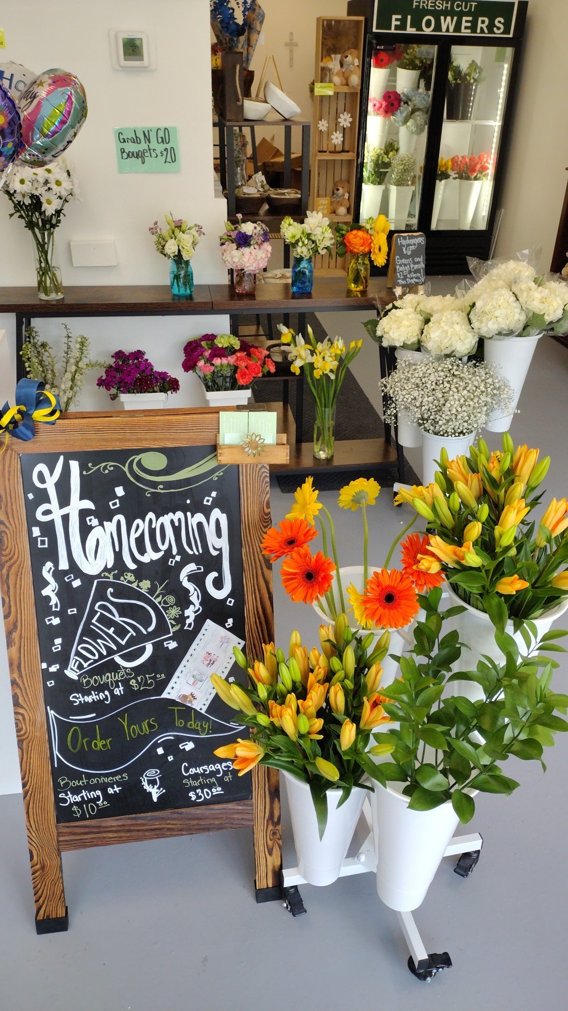 A display of fresh flowers and a blackboard sign at a flower shop. The sign promotes homecoming flowers, with prices for bouquets starting at $25, boutonnières at $10, and corsages at $30. The flowers include lilies, gerbera daisies, and other mixed blooms arranged in white vases and displayed on shelves.