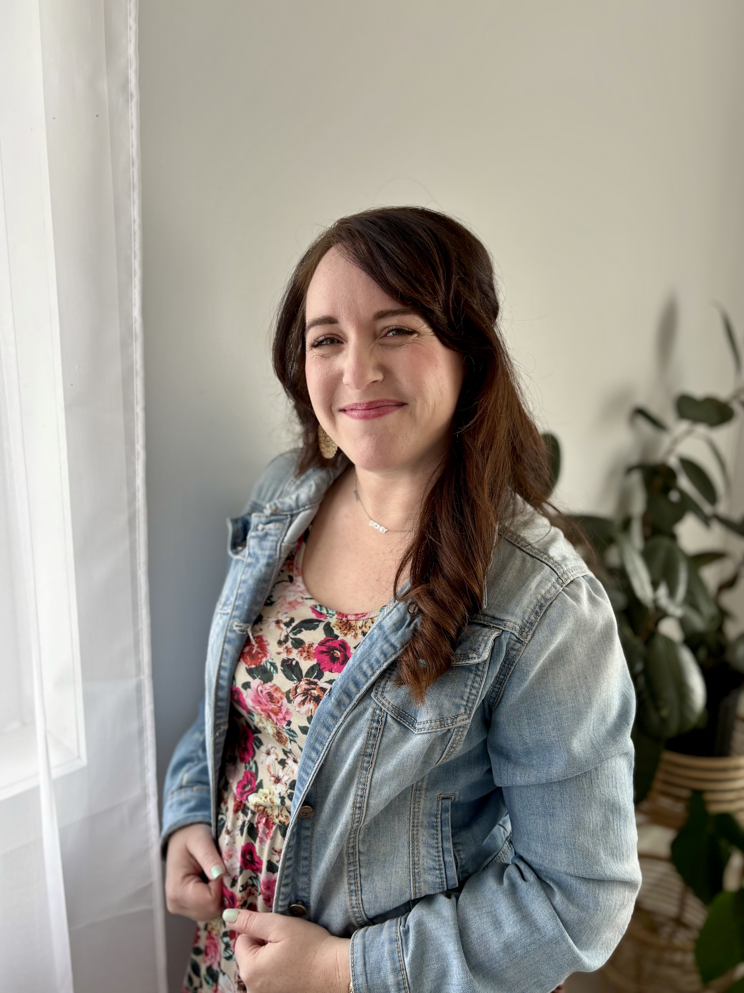 Woman smiling indoors wearing a denim jacket and floral dress standing by curtains, with a plant in the background.