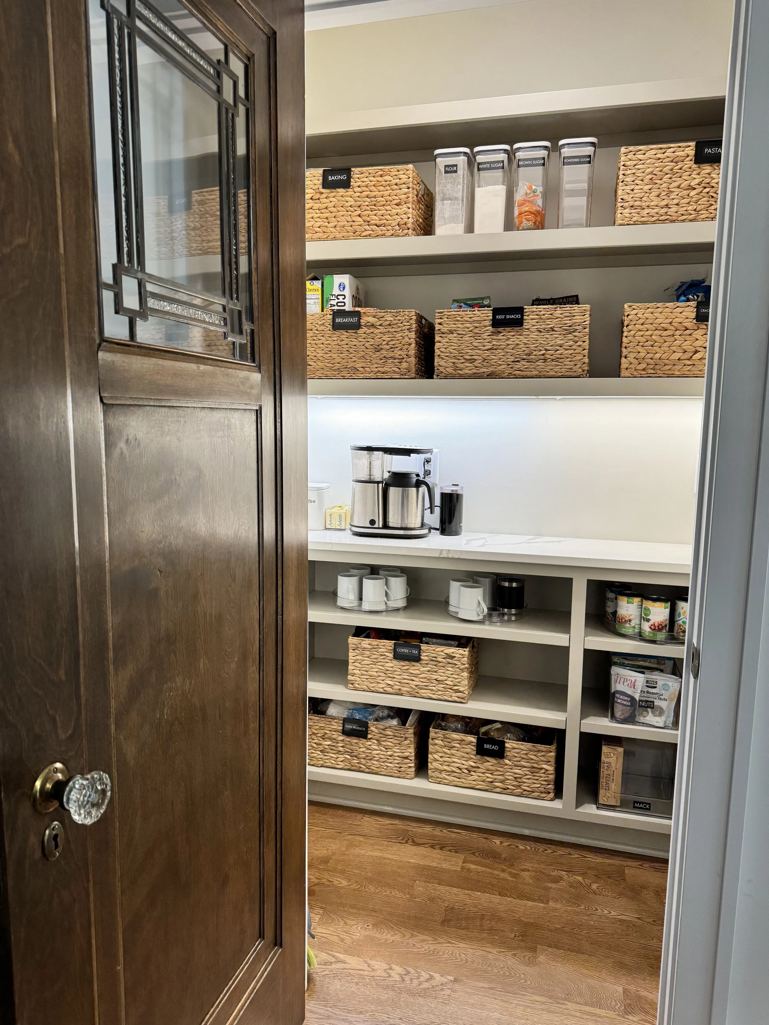 Woven baskets with black labels and clear canisters organizing food items in a pantry