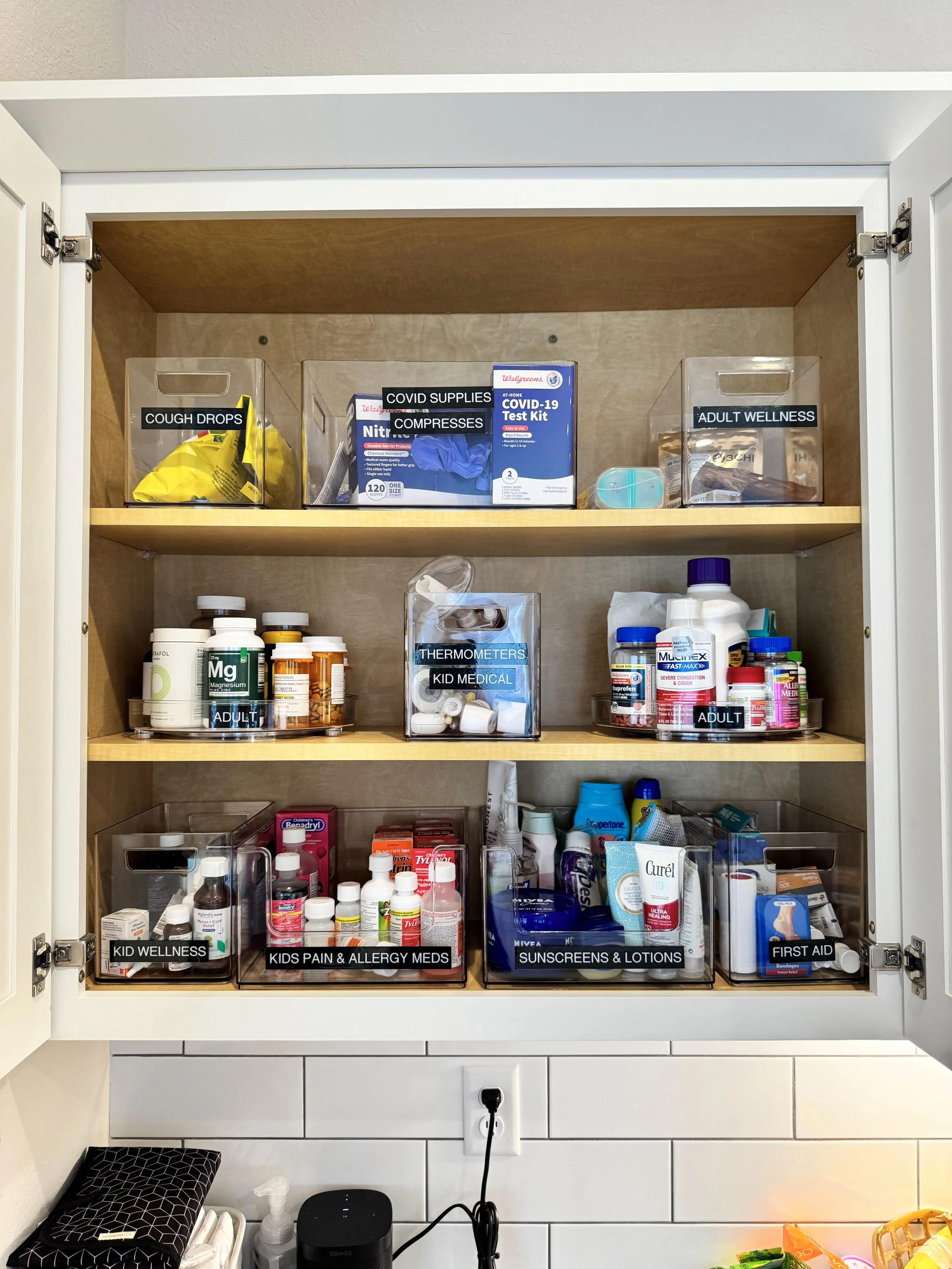 Clear containers with black labels organizing medicines and first aid inside a kitchen cabinet