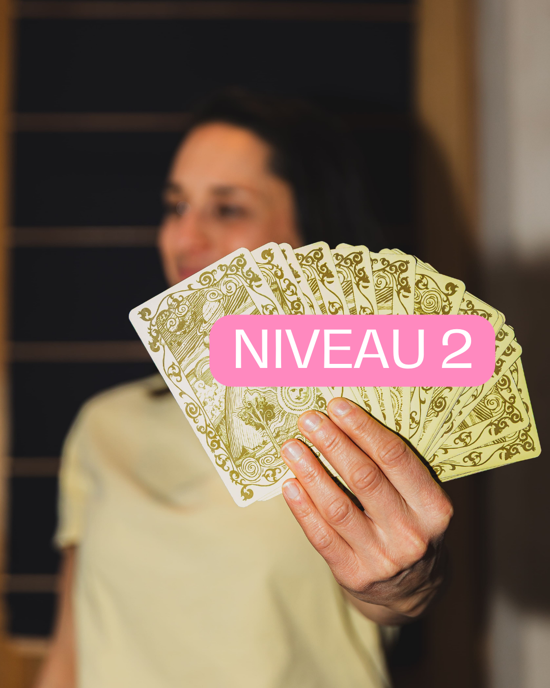 Une femme tient des cartes de tarot avec un message 'NIVEAU 2' en pink devant l'objectif, floue en arrière-plan.