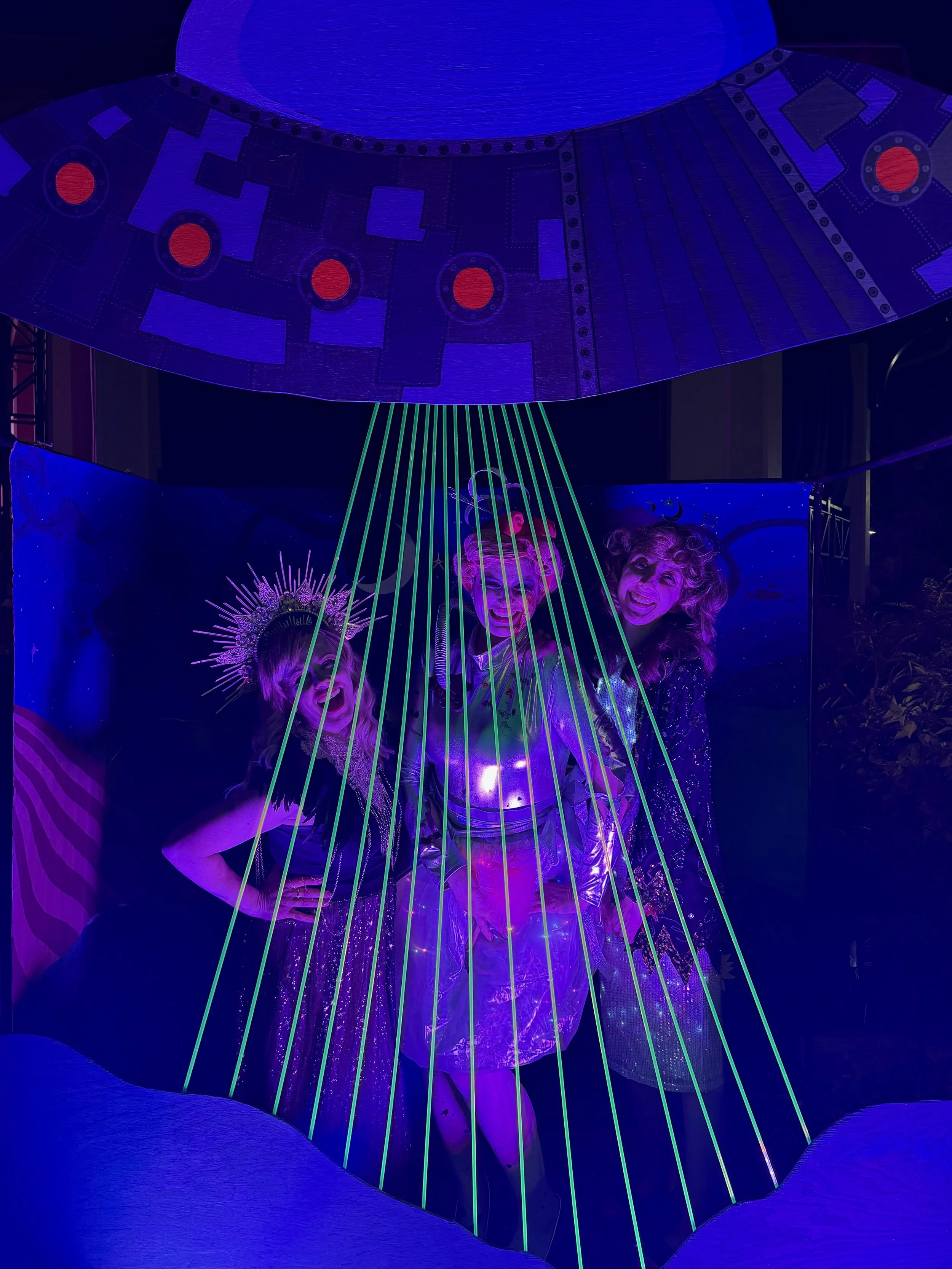 Three people dressed in space-themed costumes standing under a UFO prop emitting green light beams in a dark setting. The costumes are shiny and metallic, with accessories like a spiked headband.