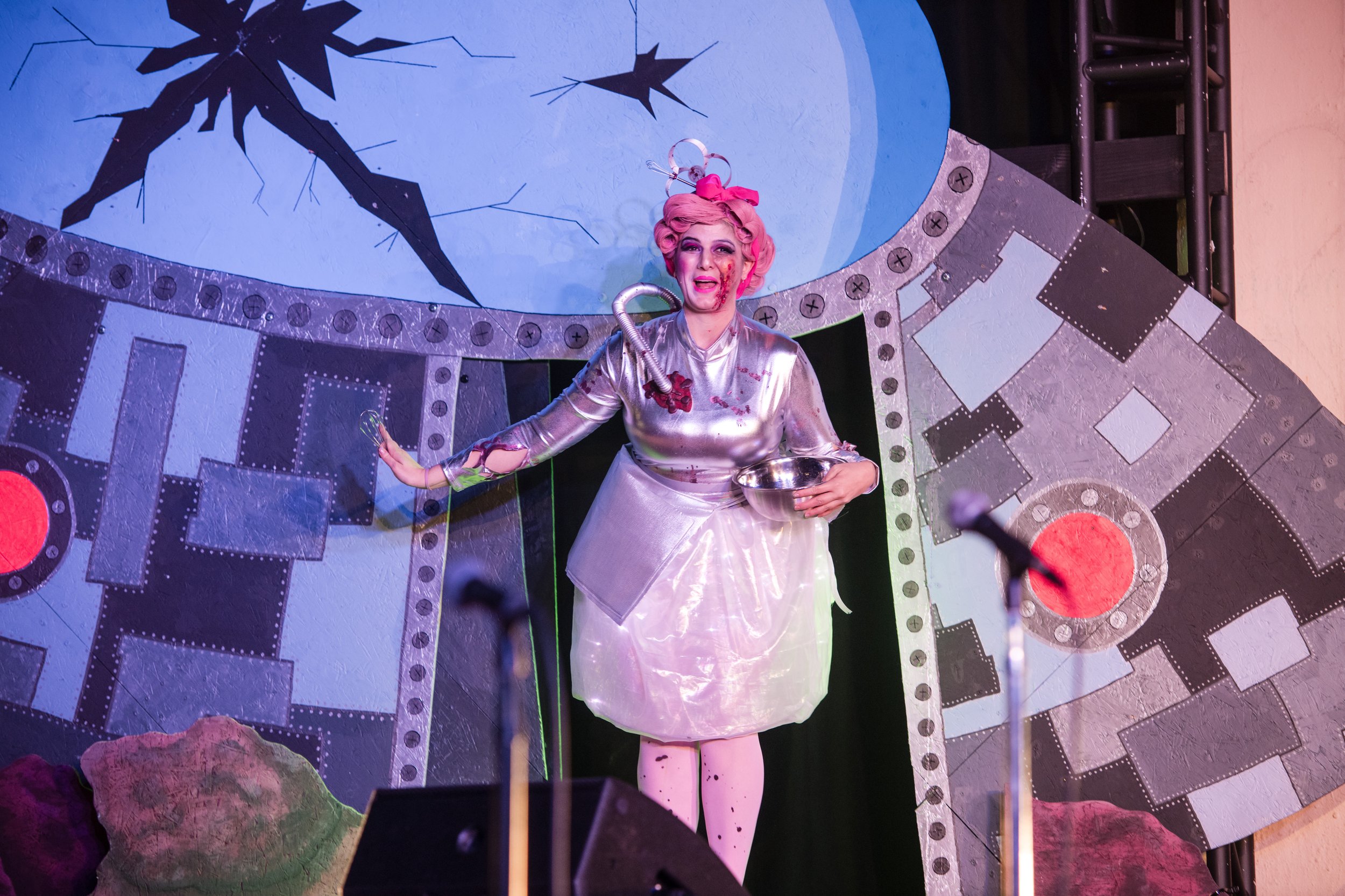 Performer in a metallic costume on stage with a sci-fi backdrop.