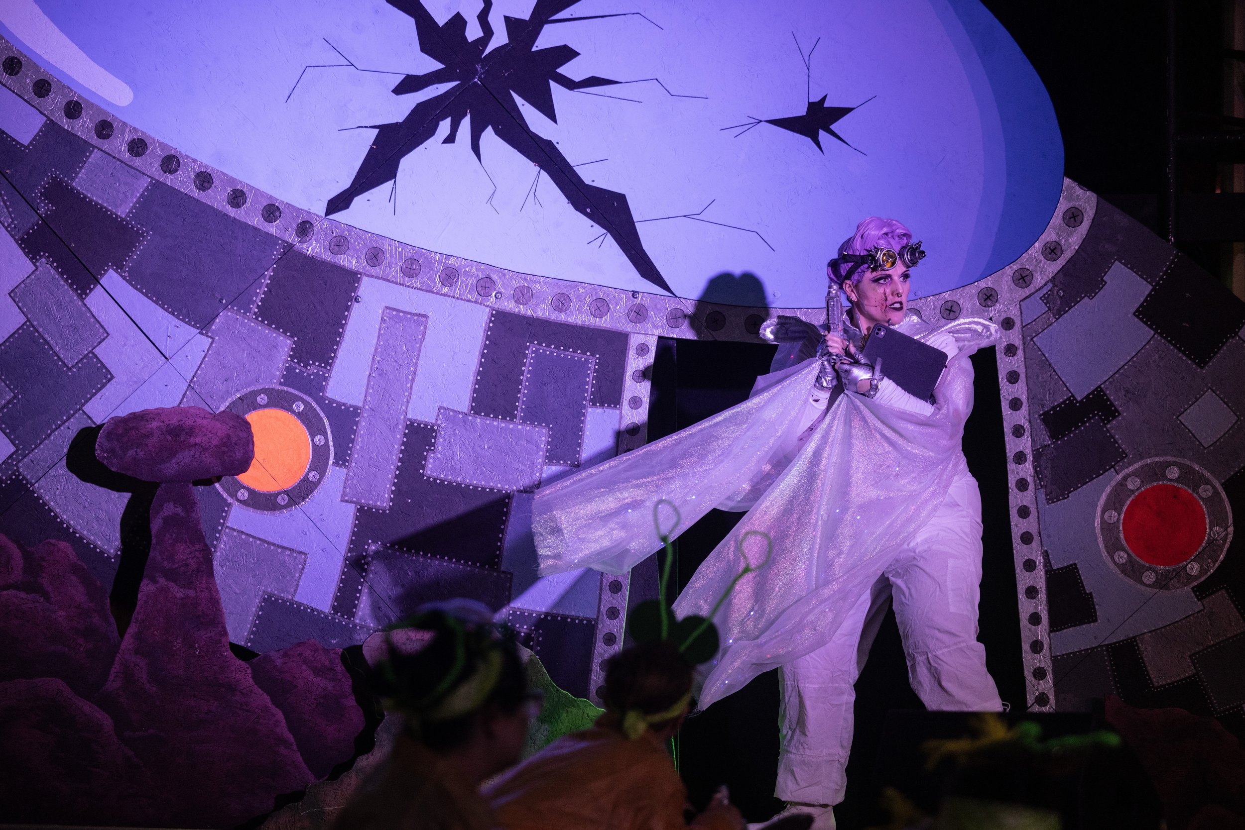 A person in a theatrical costume on stage, depicting a futuristic or sci-fi setting with a decorated backdrop. The performer is wearing white clothing, a shimmering cape, and has purple hair with goggles. The background includes a stylized broken wal