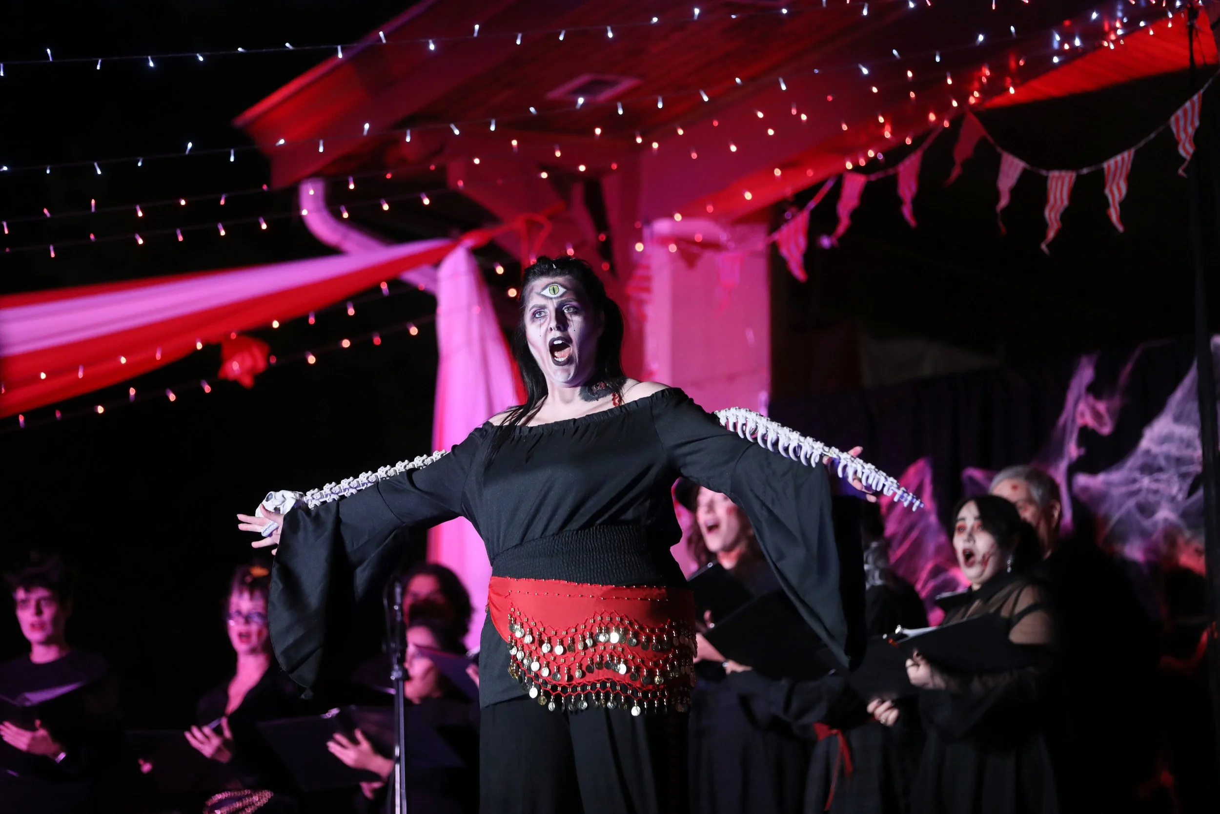 Performer in a Halloween-themed costume with a third eye painted on forehead, singing on stage under red and purple lights, accompanied by a choir in dark attire.