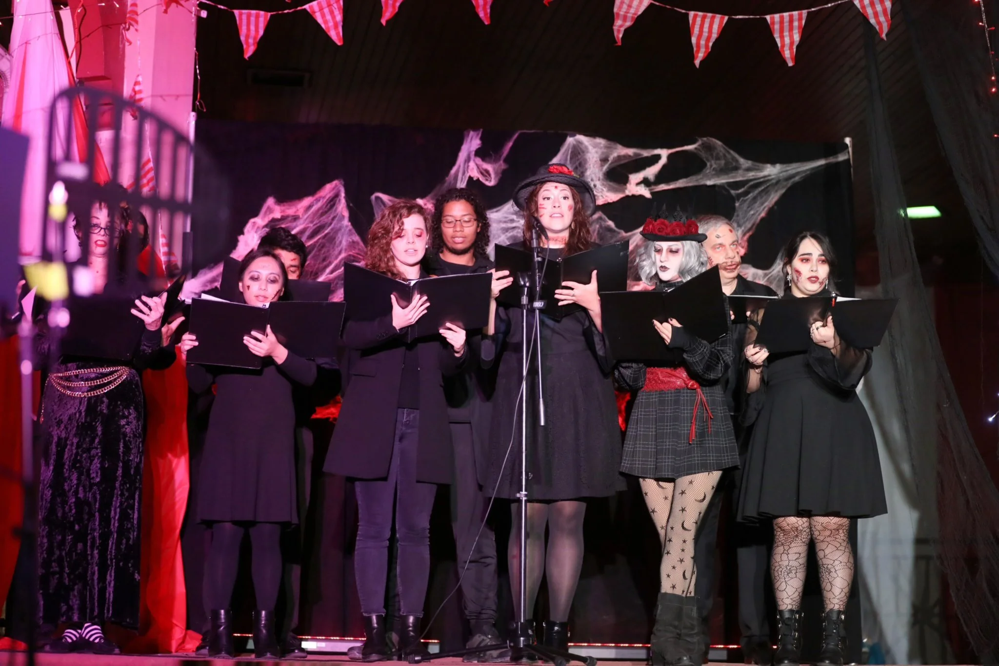 A group of people in costumes performing on stage, holding black folders, under red lighting and decorative bunting with cobwebs as a backdrop.