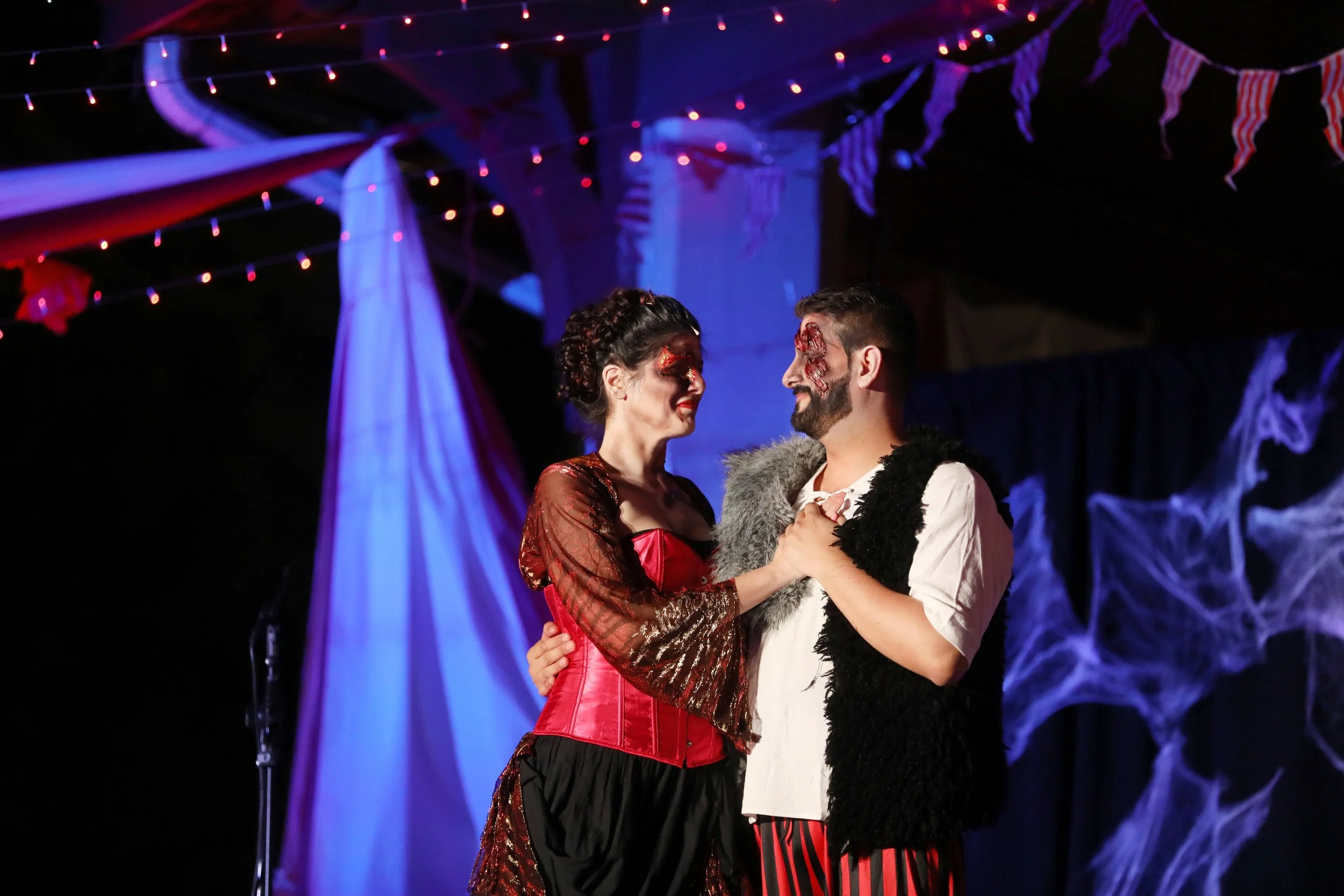 Two people in theatrical costumes with face paint, interacting on a decorated stage with purple and red lighting. The background features draped fabric and string lights.