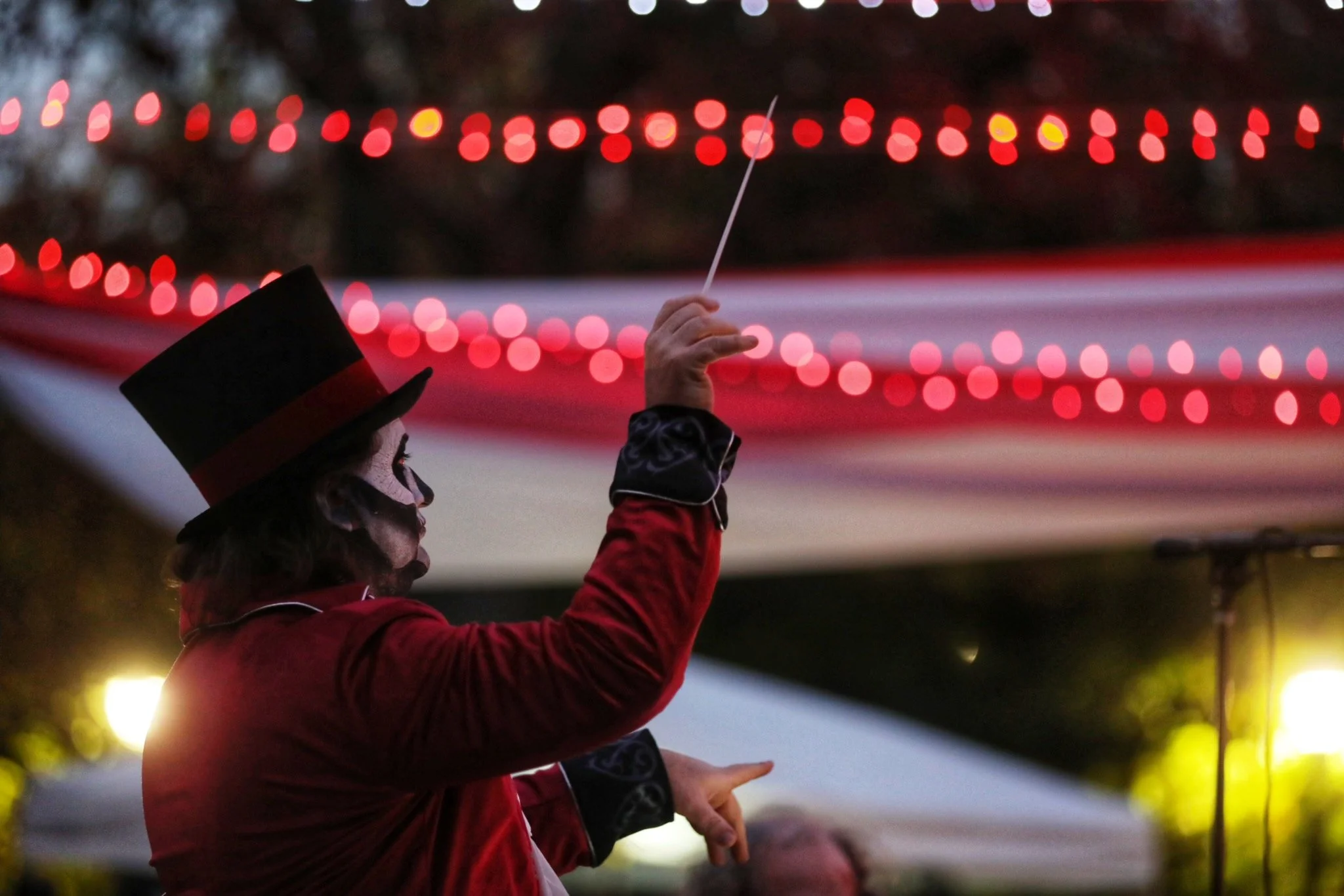 Person dressed as a circus ringmaster holding a baton with red string lights in the background.