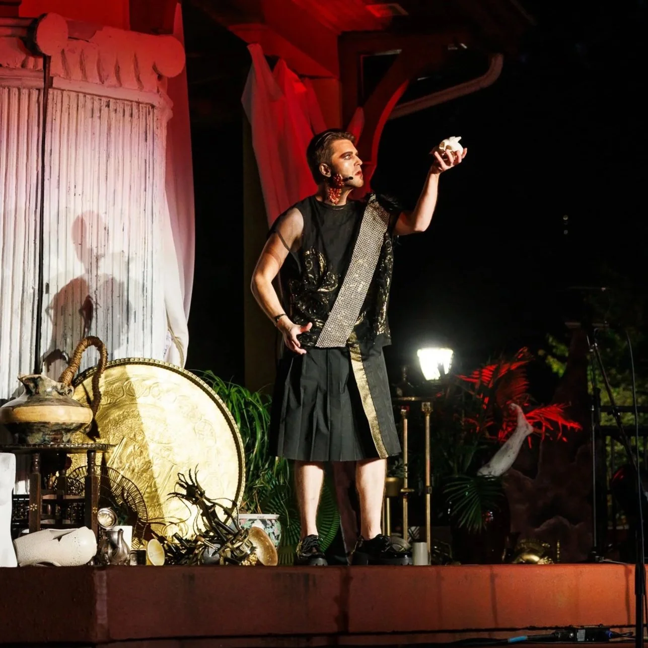 A performer in theatrical makeup and costume acts dramatically on stage with a key in his head, over a backdrop featuring a large moon with silhouettes.