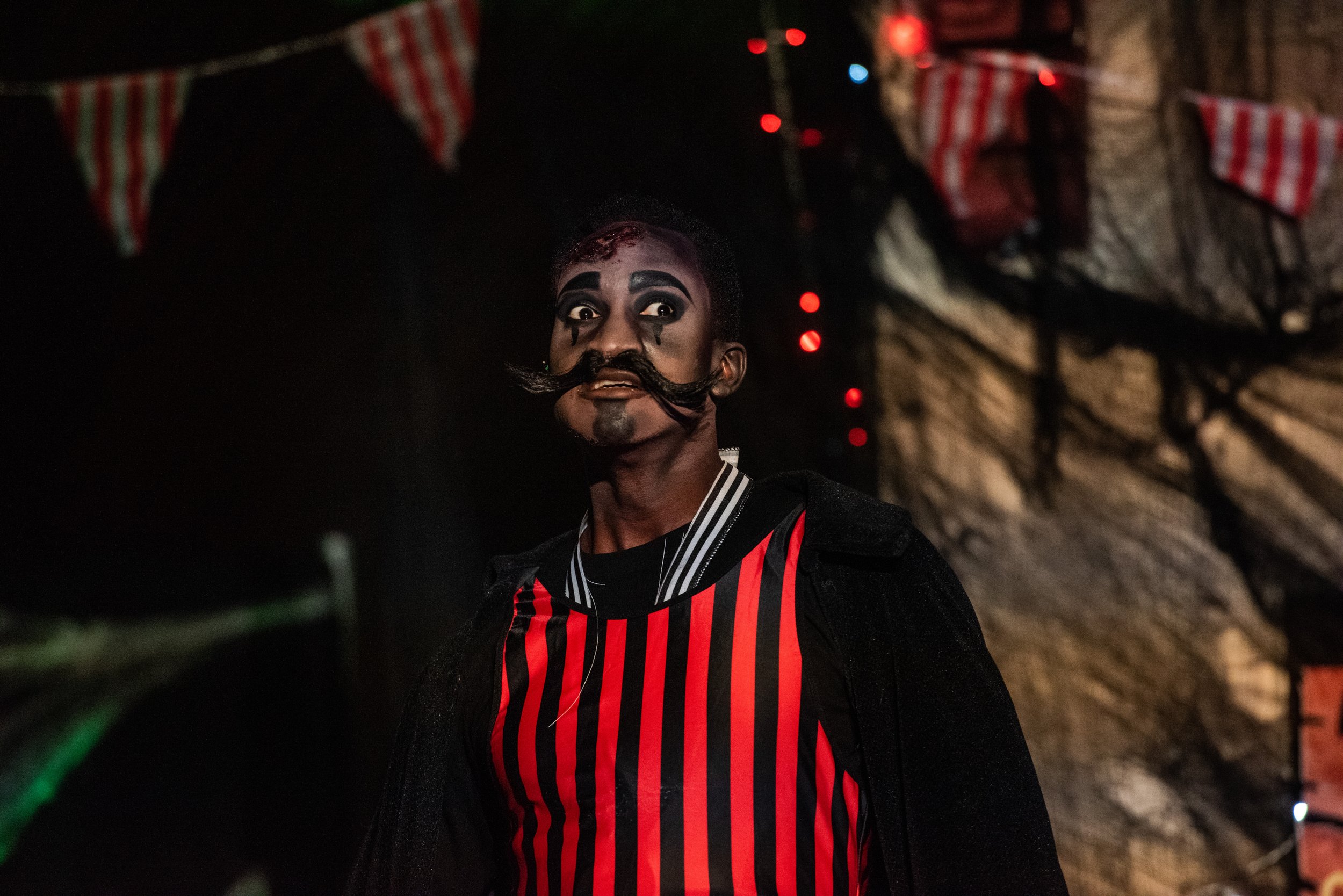 Person in circus-themed costume with red and black stripes, intricate face paint, and a theatrical mustache indoors with festive banners.