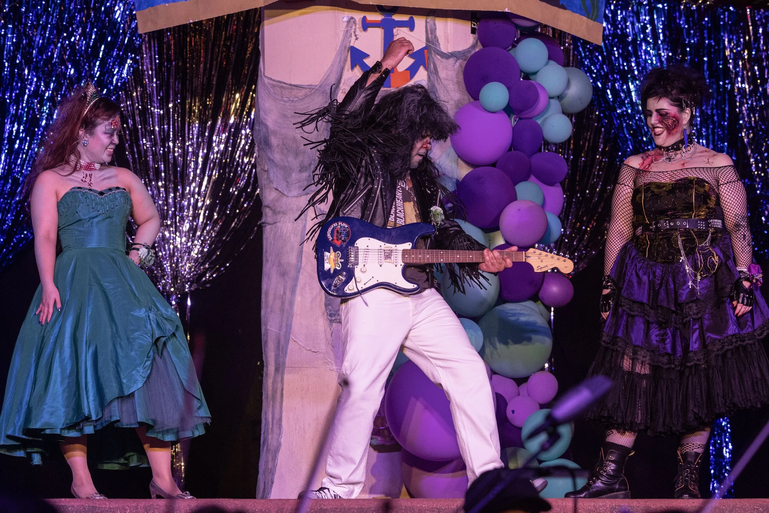 Three people in costume on stage, with one playing a blue electric guitar. One wears a green strapless dress and tiara, another in a black leather jacket with fringes and white pants, and the third in a gothic black and purple outfit. A balloon arch is in the background.