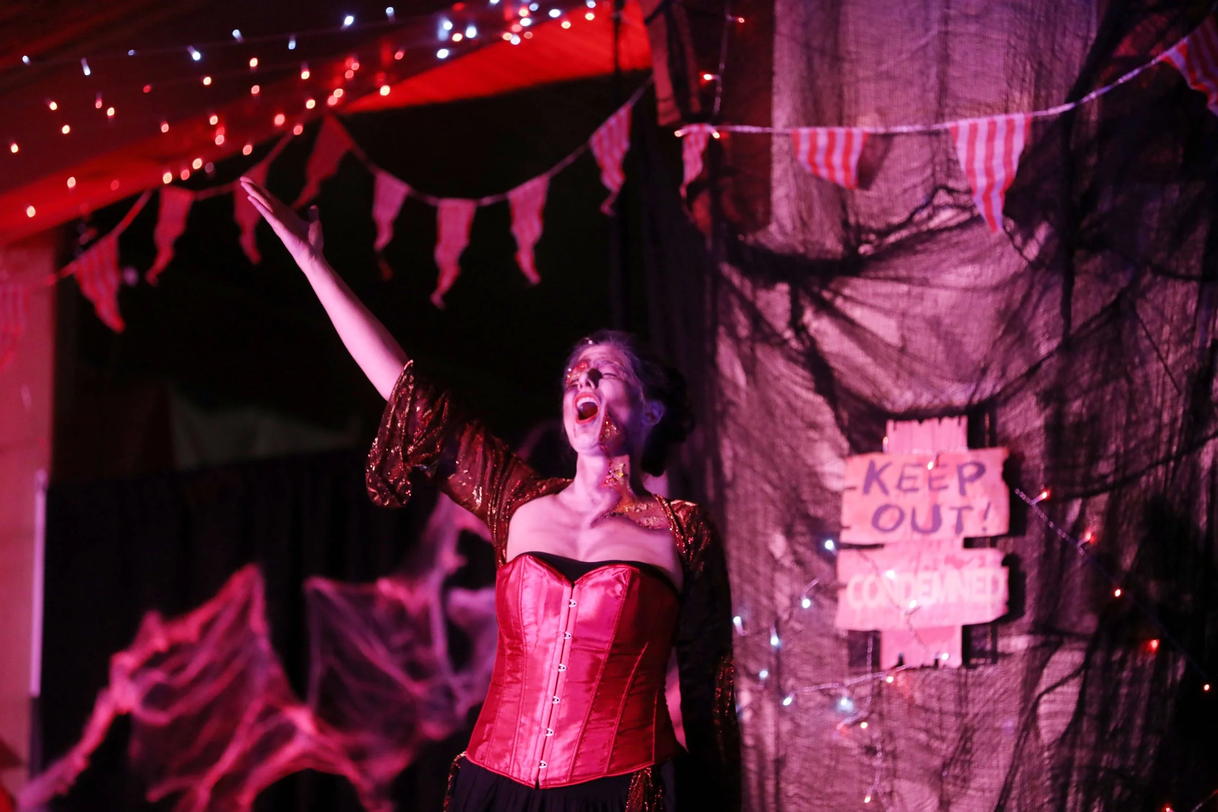 Person in a theatrical setting wearing a red corset, raising one arm, with Halloween decorations and a "Keep Out" sign in the background.