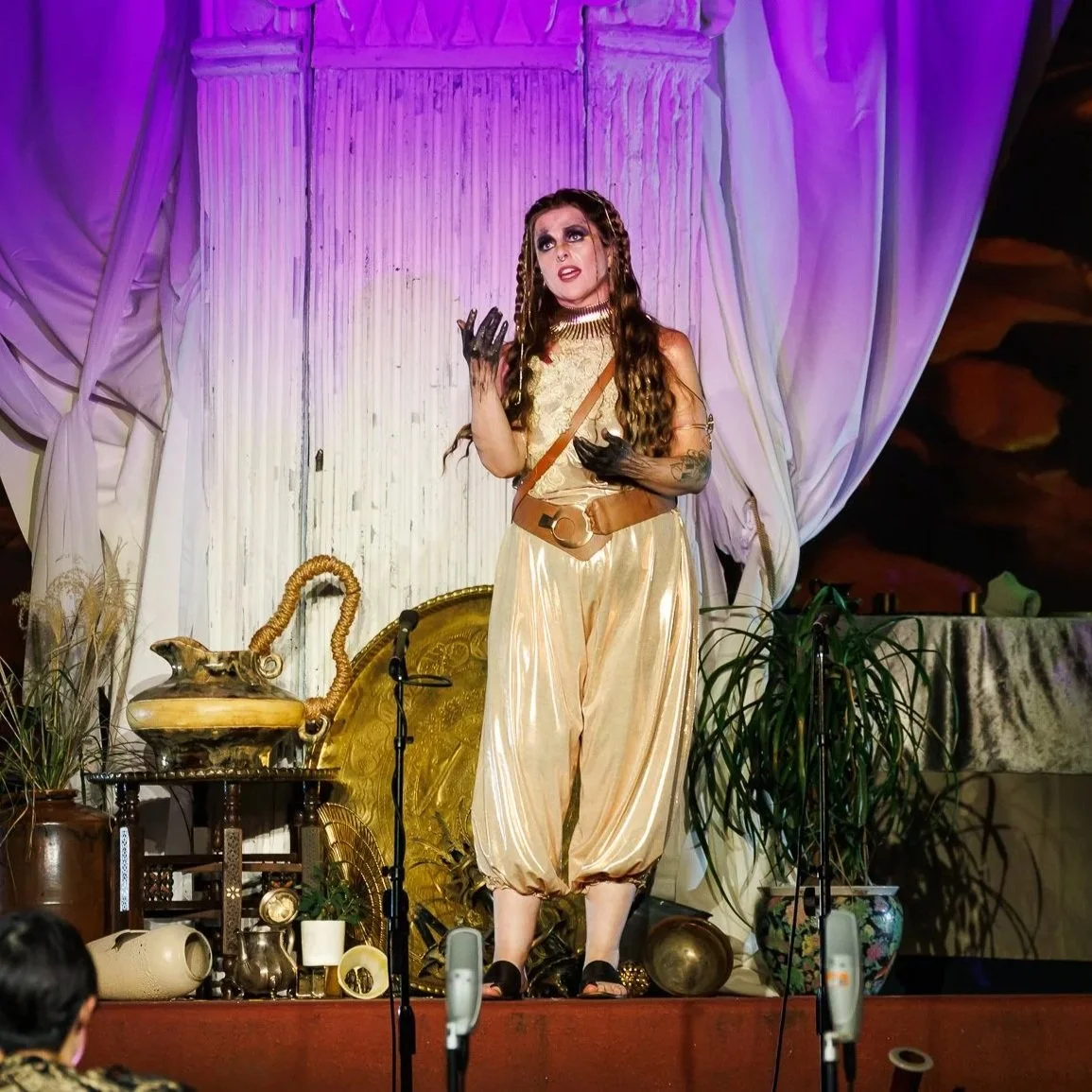 A performer dressed in a futuristic space costume with a shiny cape and goggles stands on stage against a sci-fi backdrop featuring geometric patterns and a large crack design. Bright lighting highlights the theatrical scene.