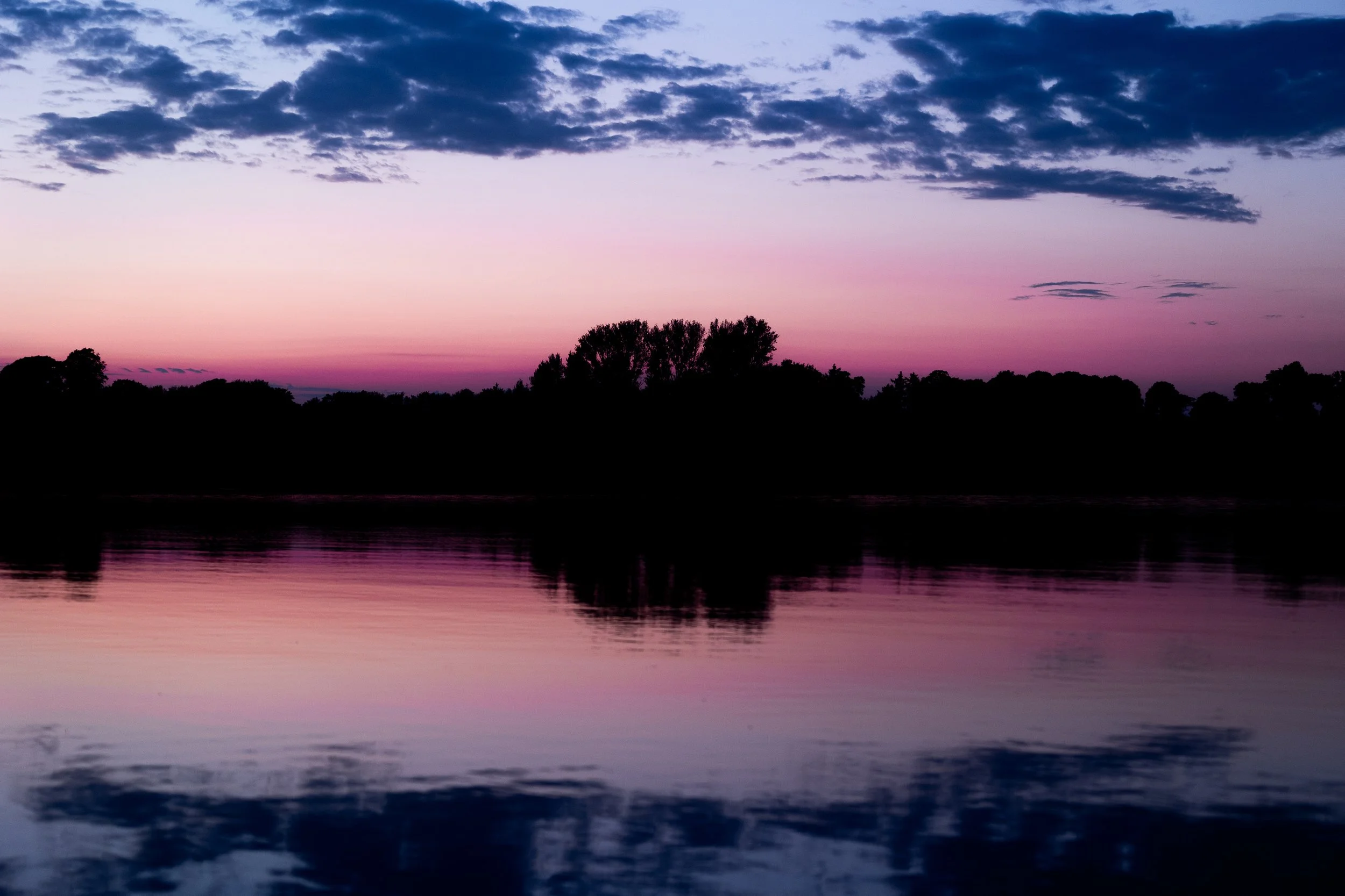 Sunset over a river with trees in silhouette and pink, purple, and blue sky with clouds.