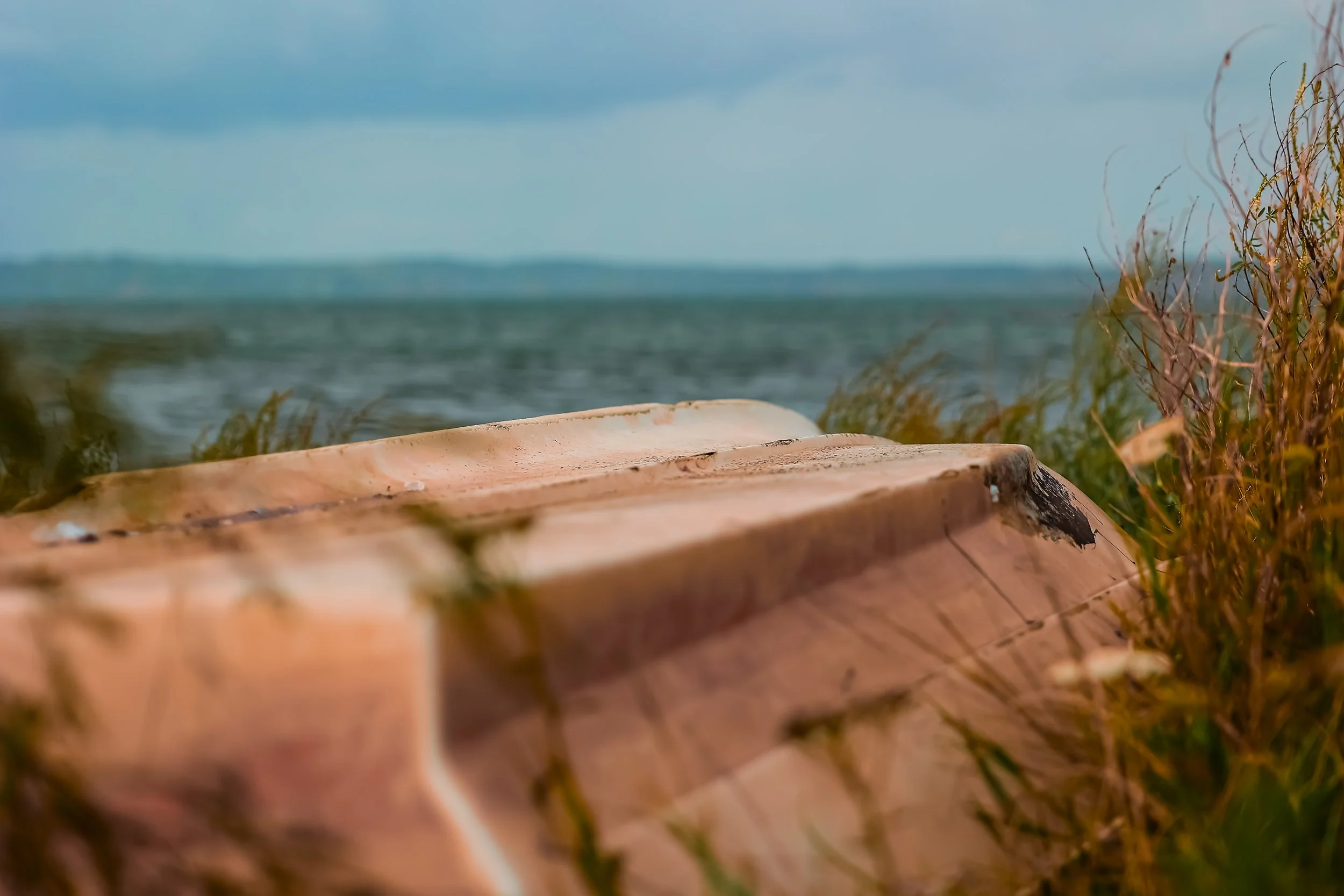 A wooden boat on the beach near water with grass and plants in the foreground and a cloudy sky in the background.