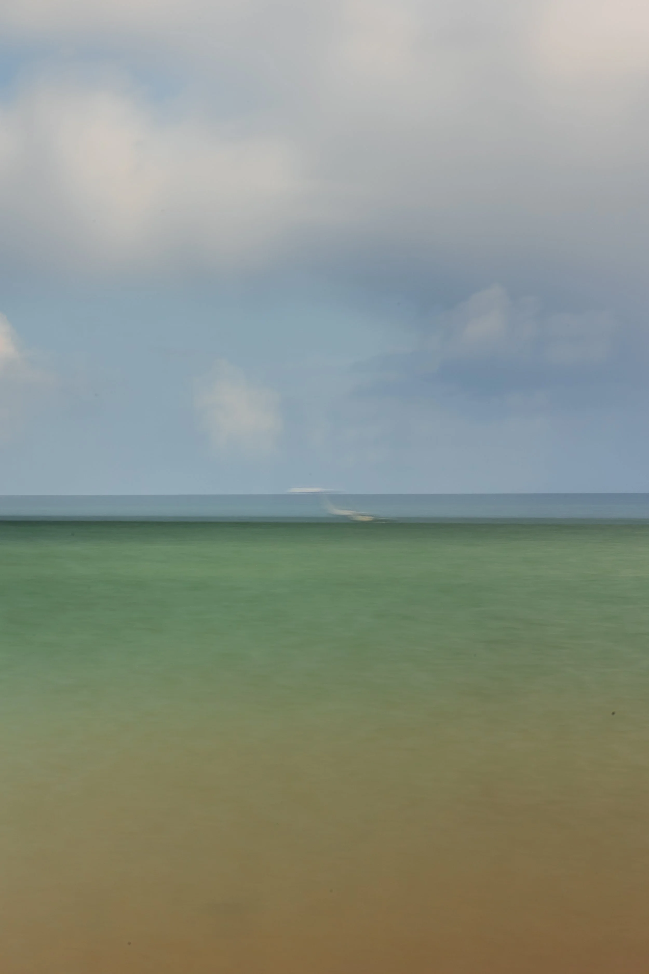 A blurry seascape with a greenish-brown shoreline, a dark blue ocean, a cloudy sky, and a distant boat kicking up a spray of water.